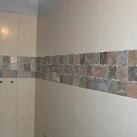 Ściana łazienki wyłożona jasnymi, beżowymi płytkami ceramicznymi z poziomym pasem dekoracyjnym z mozaiki w odcieniach szarości, brązu i beżu.