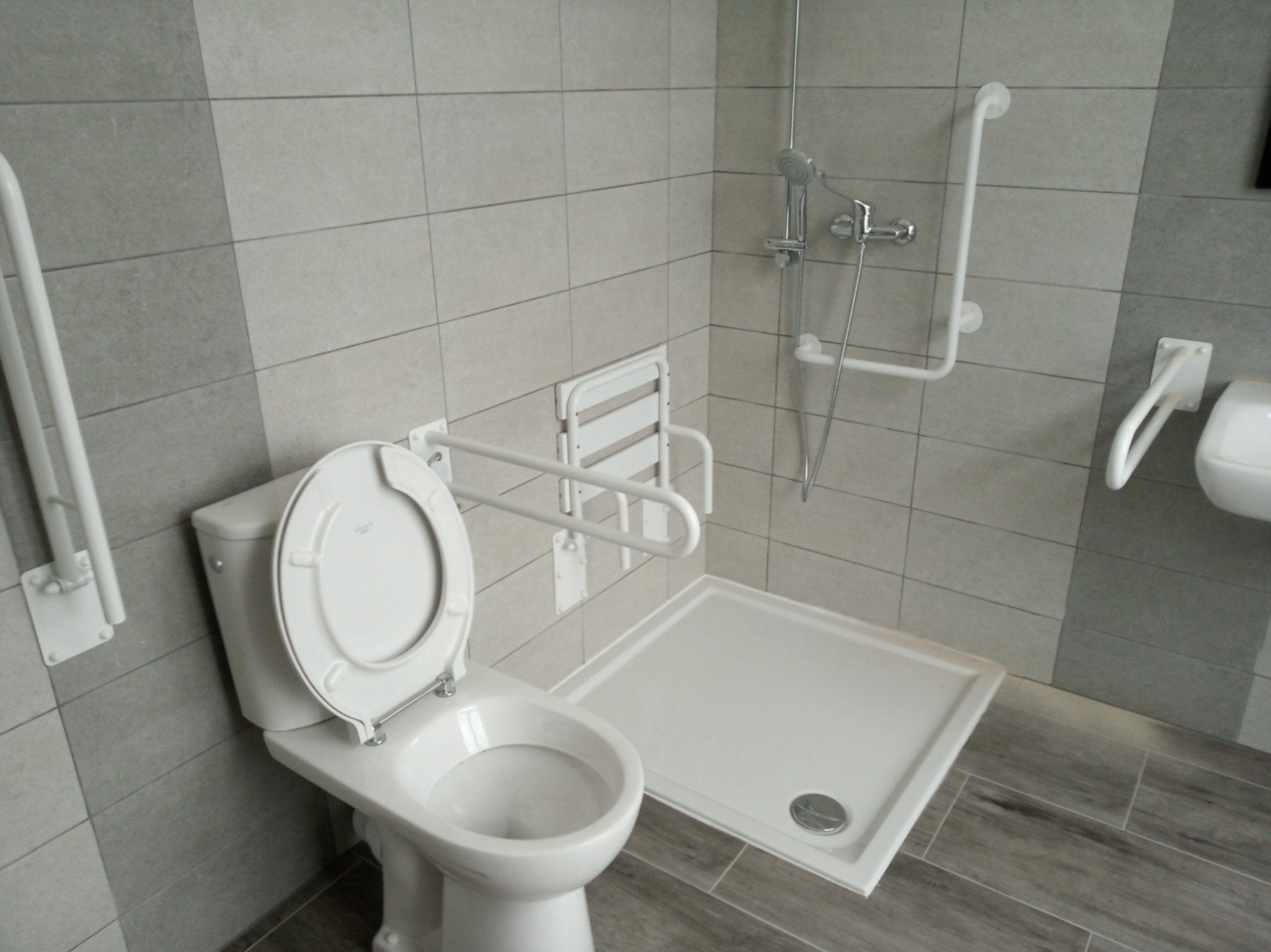 Jasna łazienka z udogodnieniami dla osób niepełnosprawnych: toaleta z uchylnymi poręczami, składane siedzisko prysznicowe i brodzik z niskim progiem.