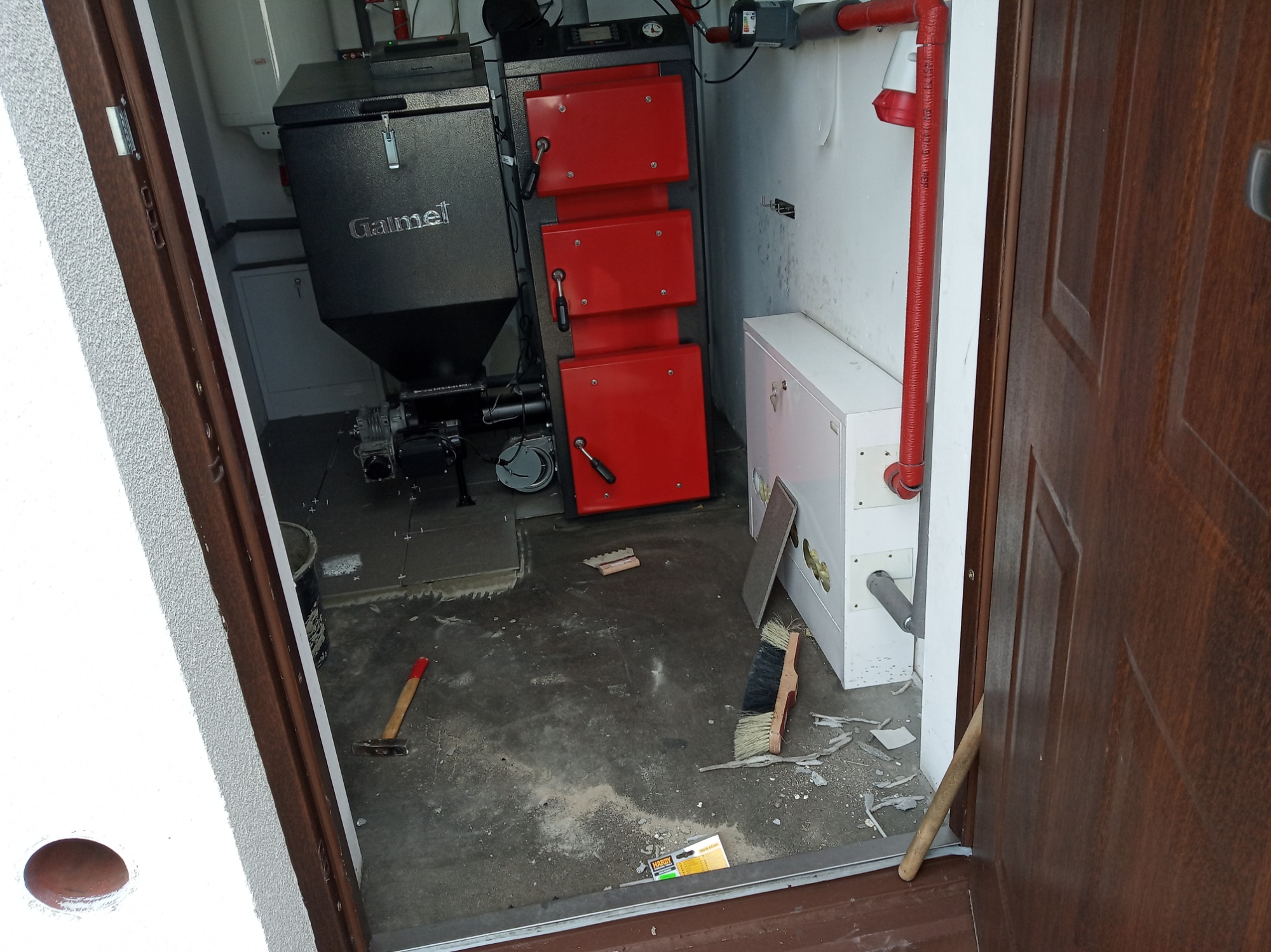 Kotłownia w trakcie prac: piec Galmet, czerwona instalacja rurowa, narzędzia glazurnicze i młotek na podłodze, widok z progu drzwi.