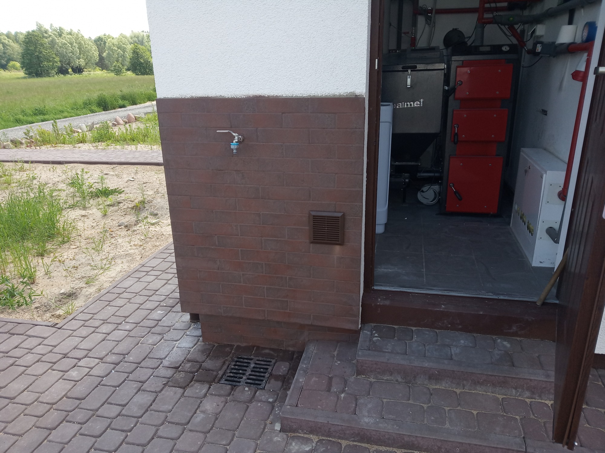 Kotłownia z piecem na paliwo stałe widoczna z zewnątrz budynku, z brązową cegłą na elewacji i kostką brukową na podłożu.