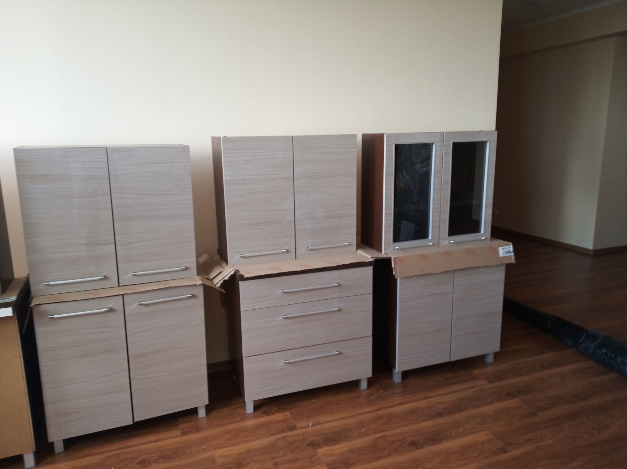 Nierozpakowane, jasne meble modułowe z drewnopodobnym wzorem, ustawione na podłodze w trakcie montażu, zabezpieczone kartonem.