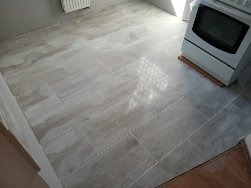 Ułożona podłoga z płytek imitujących marmur, widoczne wąskie fugi i fragment kuchenki gazowej w rogu pomieszczenia.