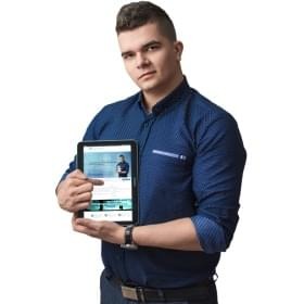 Młody mężczyzna w niebieskiej koszuli prezentuje tablet z widoczną stroną internetową i wskazuje na nią palcem. Na nadgarstku widoczny zegarek. Tło białe.