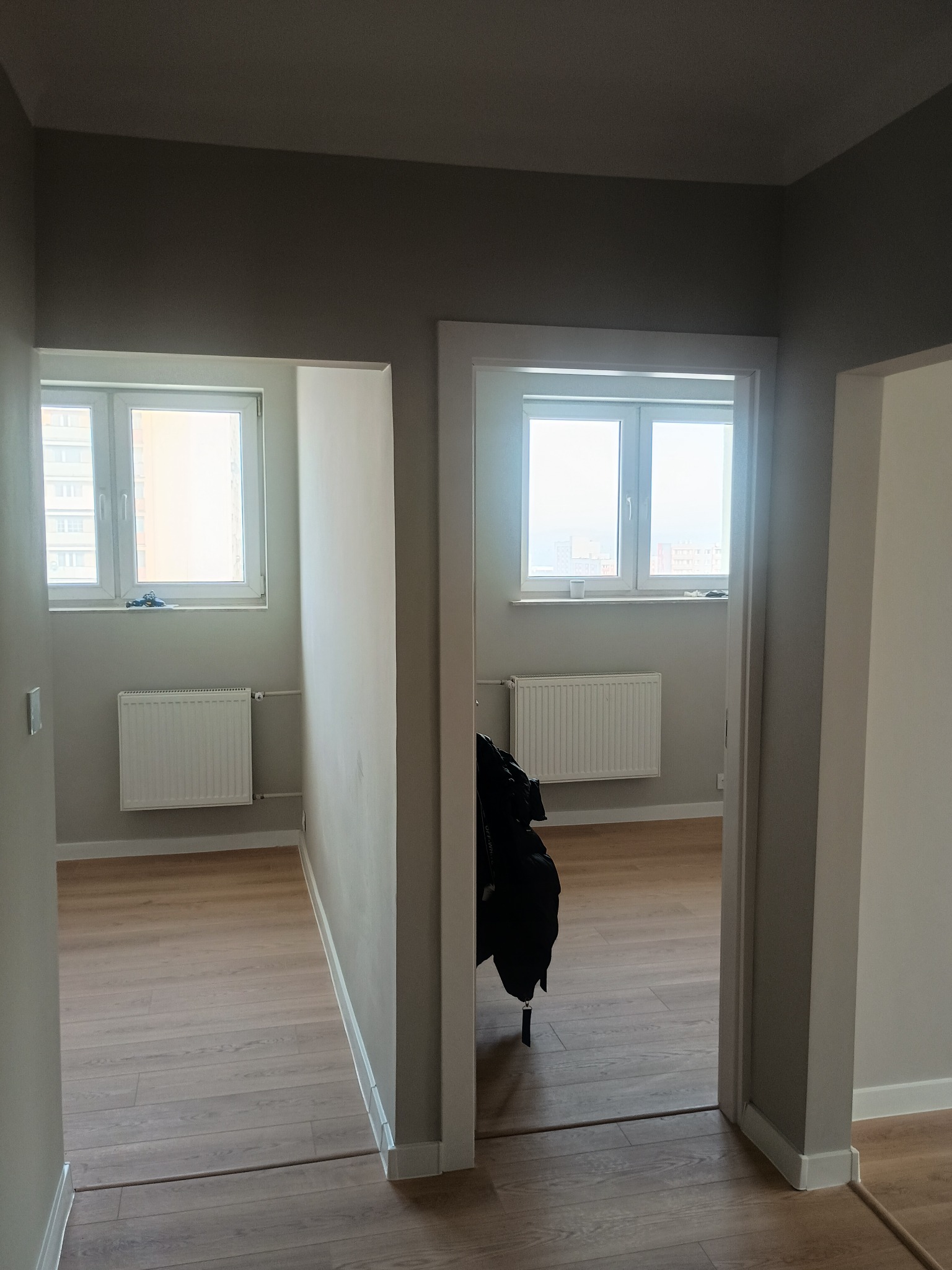 Jasne wnętrze po remoncie: świeżo pomalowane ściany w odcieniach szarości, ułożone panele podłogowe, białe grzejniki pod oknami. Widok z korytarza do pokoju.