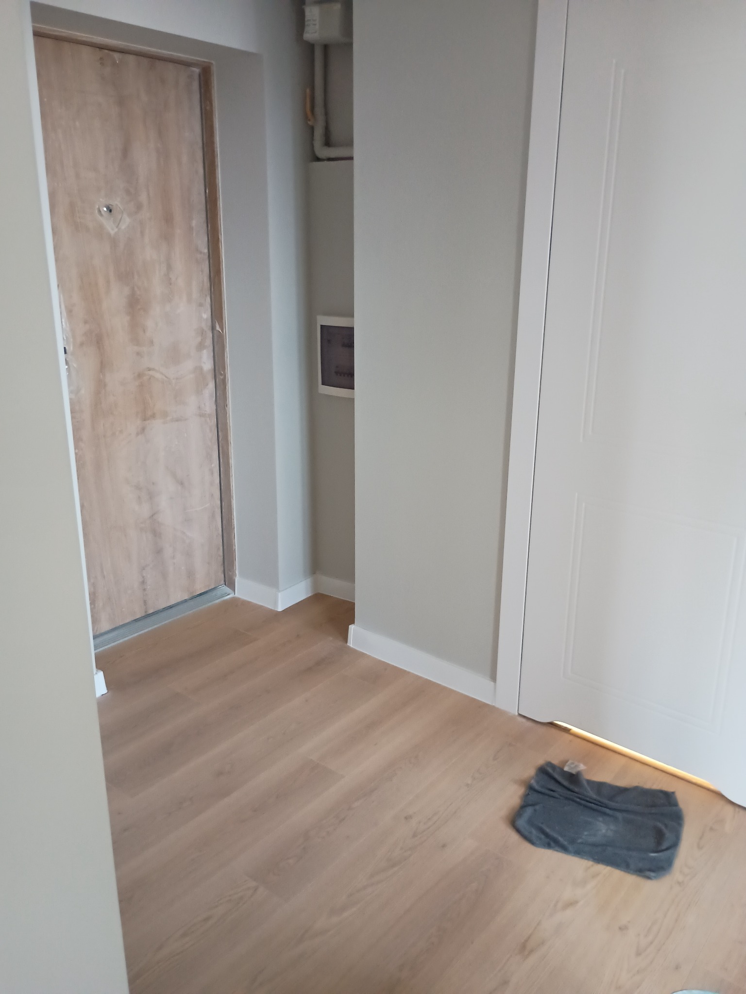Jasne panele podłogowe w korytarzu po remoncie, widoczne drzwi wejściowe i wewnętrzne, ściany pomalowane na szaro, listwy przypodłogowe białe, szmatka na podłodze.