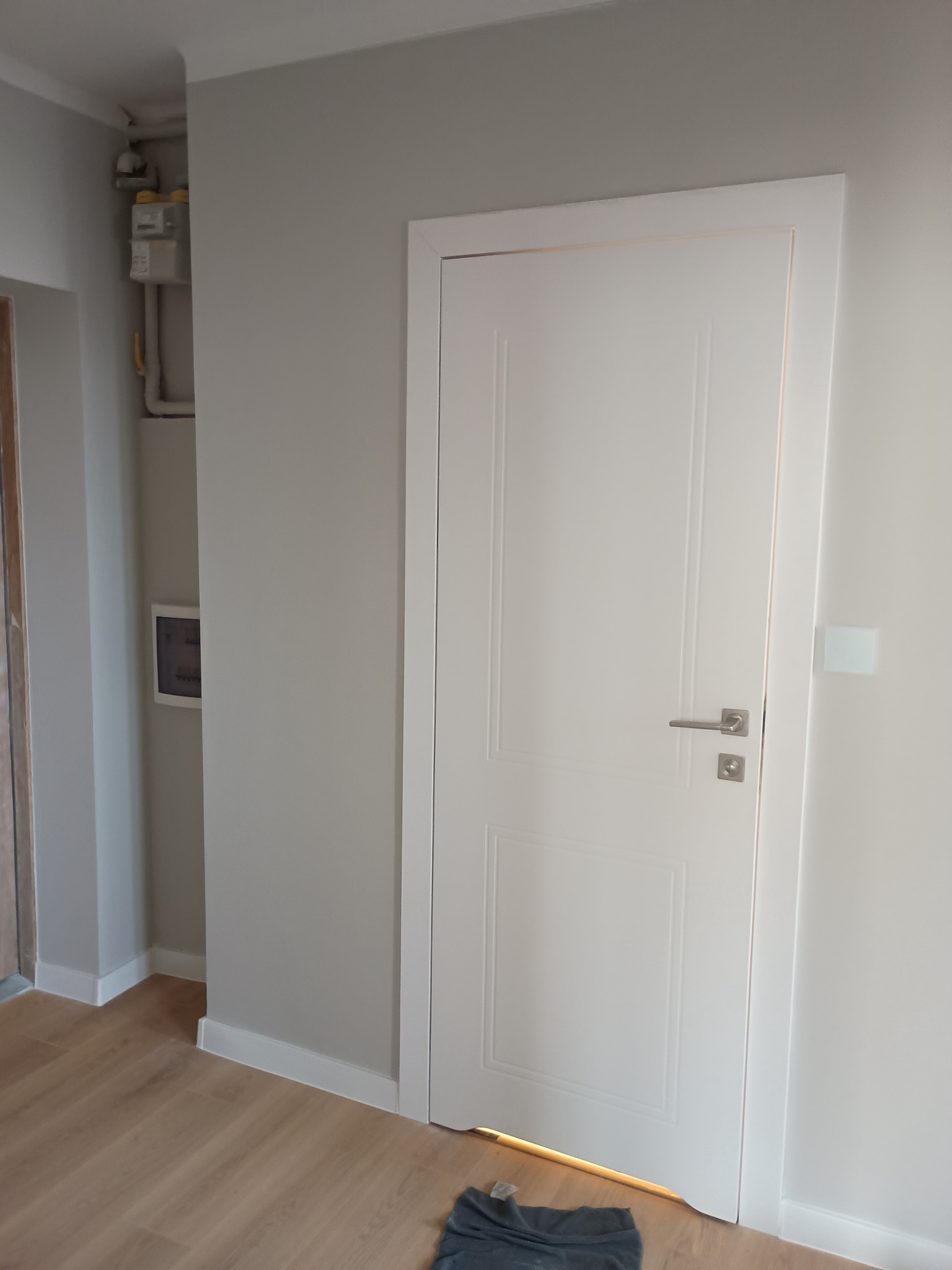 Białe, frezowane drzwi wewnętrzne z srebrną klamką, osadzone w ościeżnicy. Ściany pomalowane na szaro, widoczna listwa przypodłogowa i fragment podłogi z paneli.