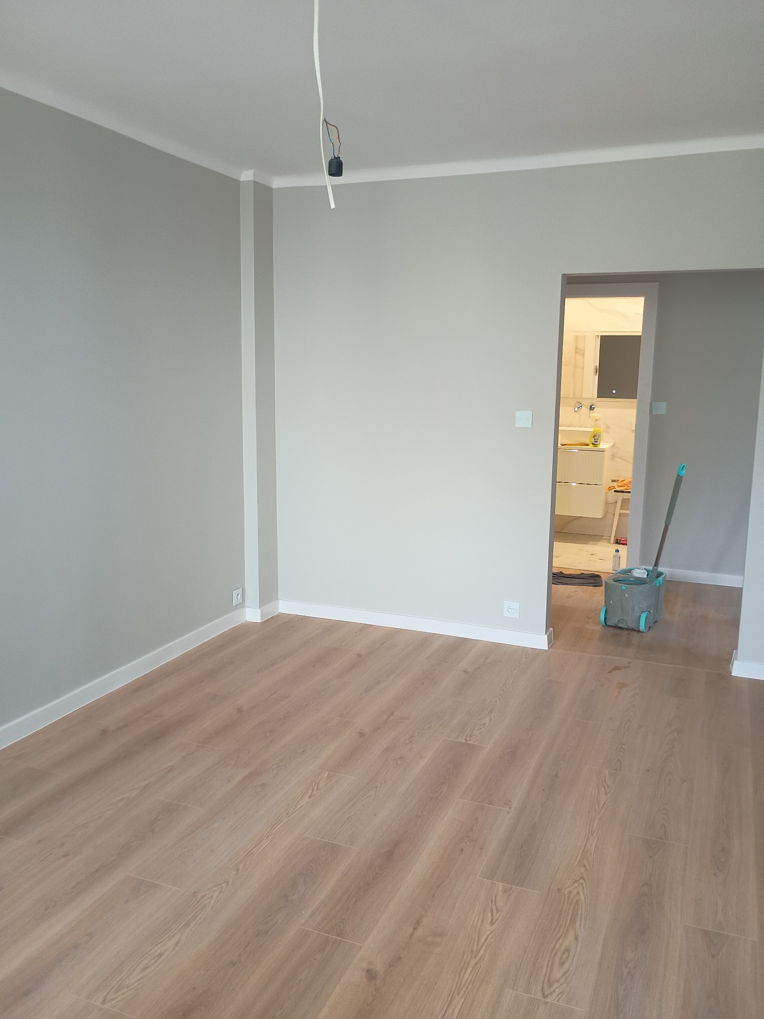 Jasnoszary pokój po remoncie z panelami podłogowymi i widokiem na łazienkę. W rogu stoi wiadro z mopem. Wiszące kable elektryczne.