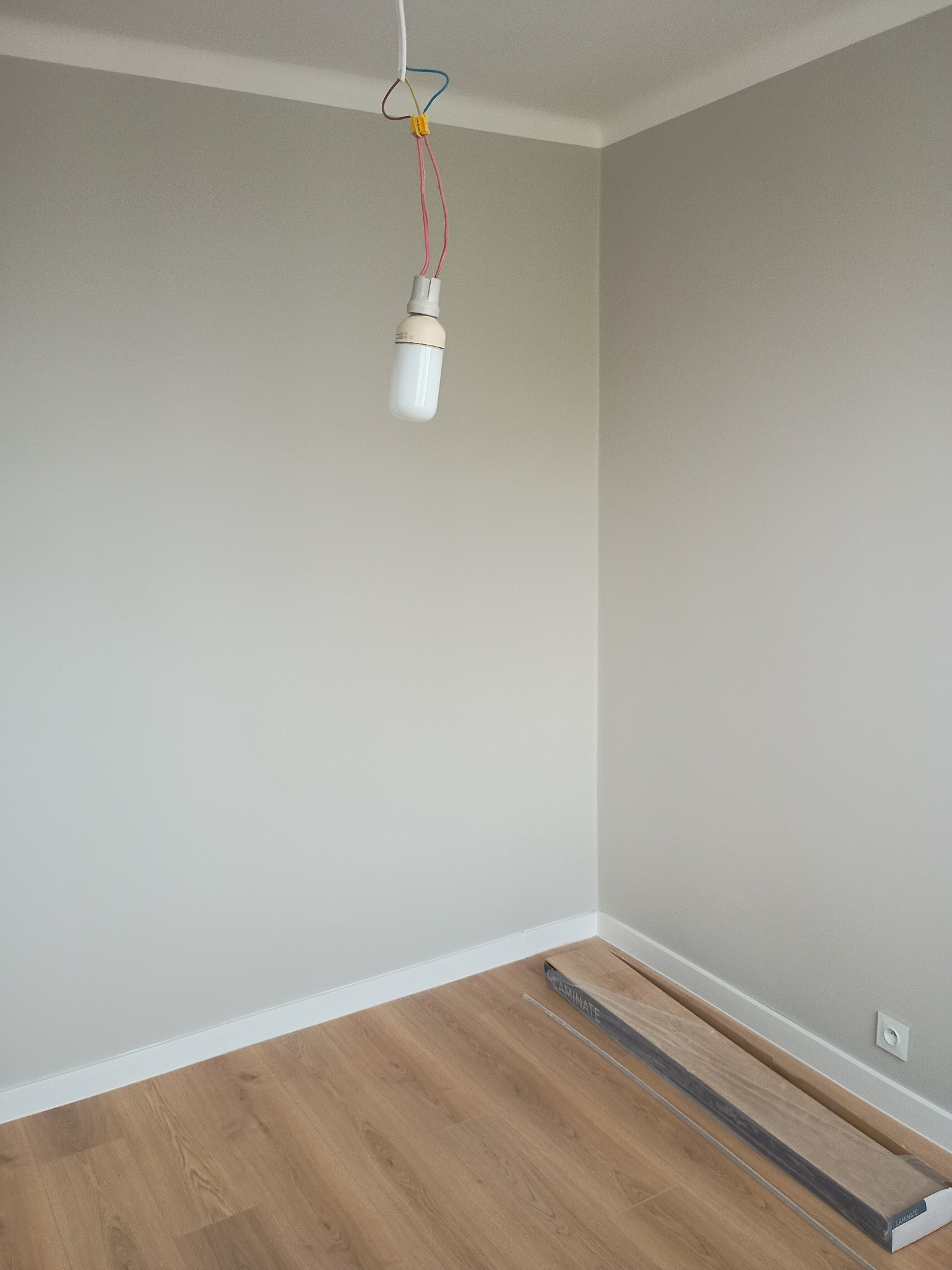 Pokój po malowaniu ścian na szaro, z ułożoną podłogą z paneli. Na podłodze leżą panele w opakowaniu, a z sufitu zwisa żarówka na kablu.