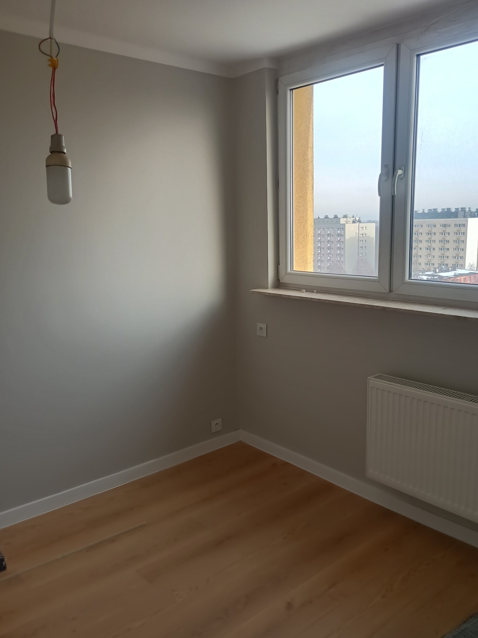 Pokój po remoncie: szare ściany, jasne panele podłogowe, białe listwy przypodłogowe i okno z widokiem na miasto. Wisząca żarówka na kablu dodaje surowego charakteru.