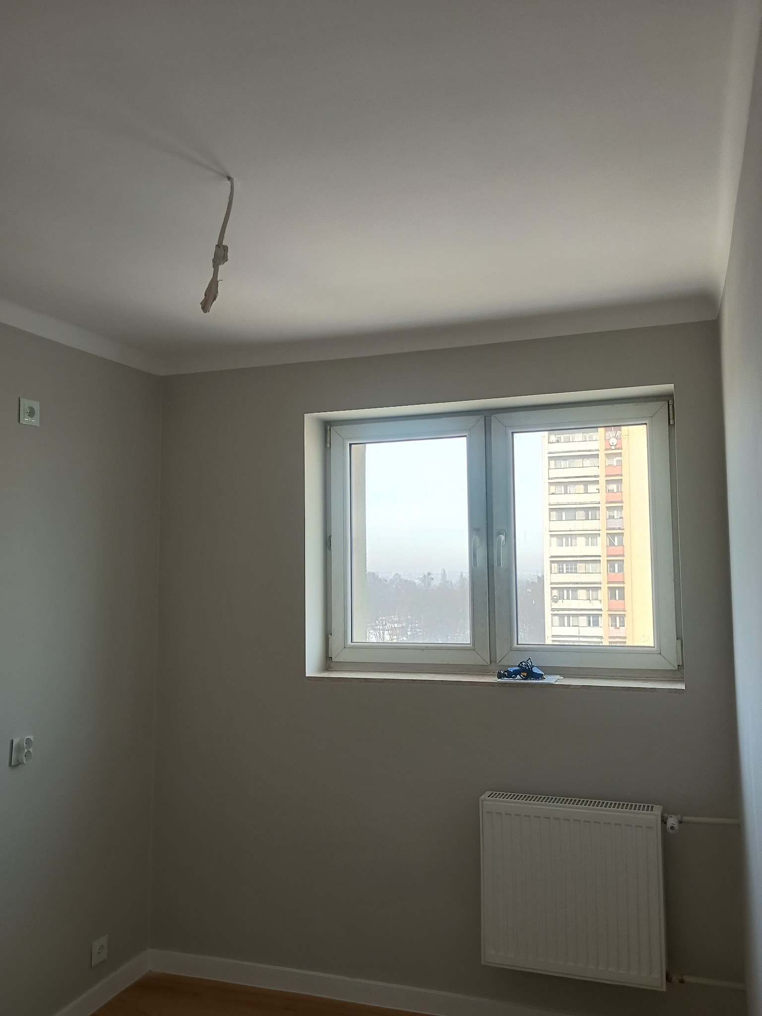 Pokój po malowaniu na szaro z białymi listwami, widok na blok przez okno. Kabel zwisa z sufitu. Grzejnik pod oknem. Gładkie ściany, świeżo po remoncie.