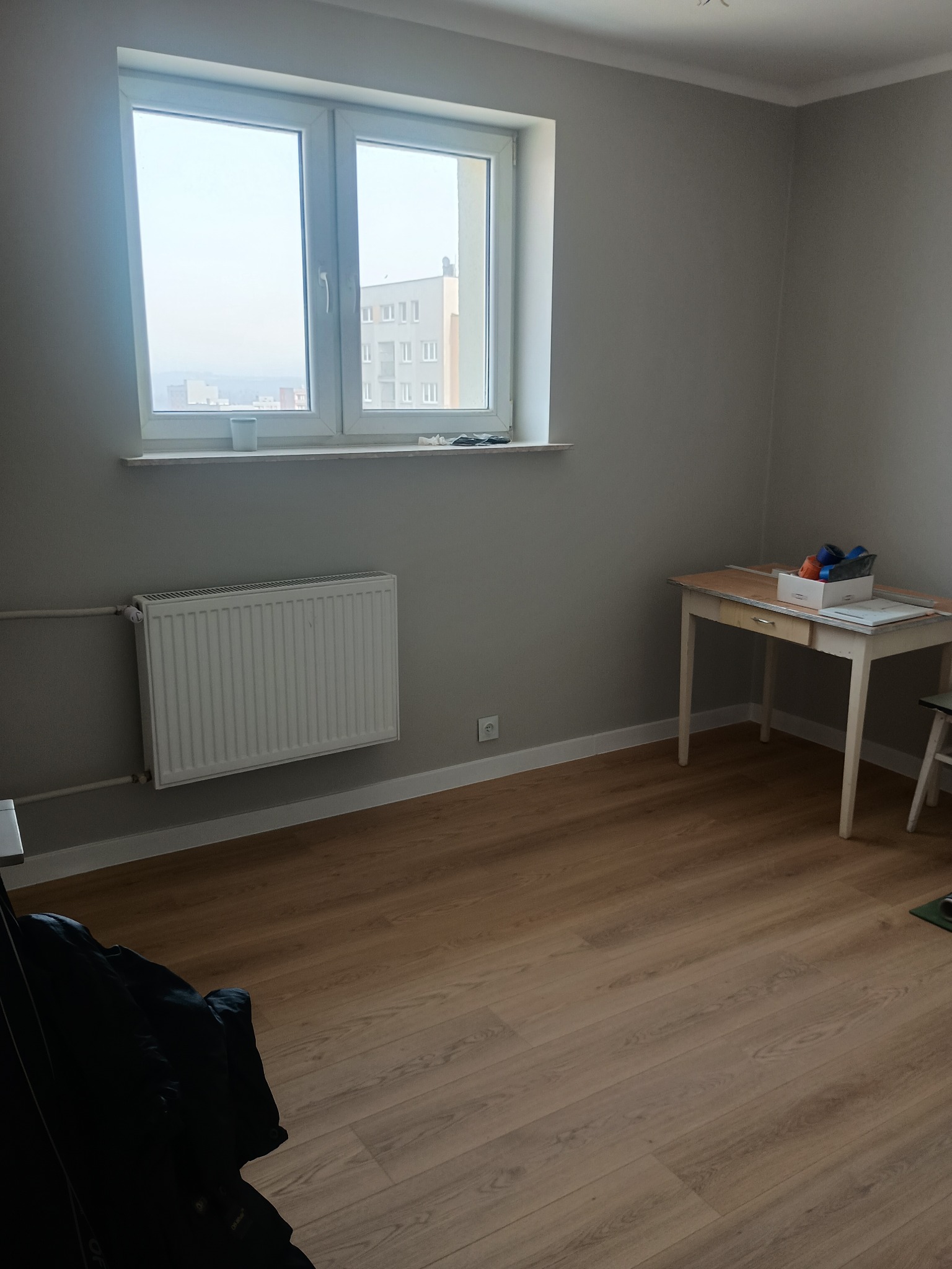 Jasne wnętrze pokoju po remoncie: świeżo pomalowane szare ściany, nowe panele podłogowe, białe okno z widokiem na miasto. Na biurku akcesoria malarskie.