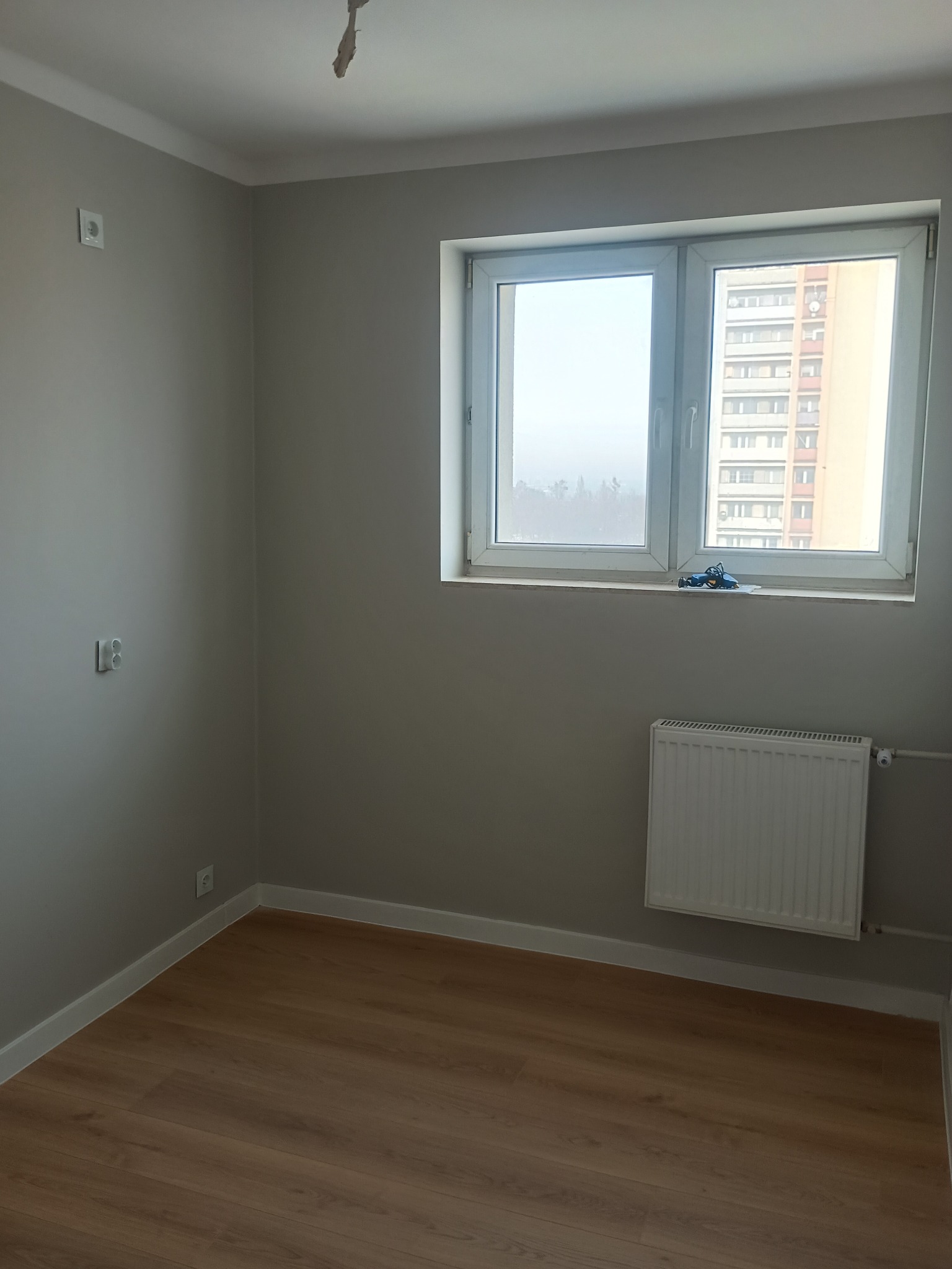 Pomalowane na szaro wnętrze pokoju z panelami podłogowymi, białym grzejnikiem i oknem z widokiem na blok. Na parapecie leżą narzędzia.