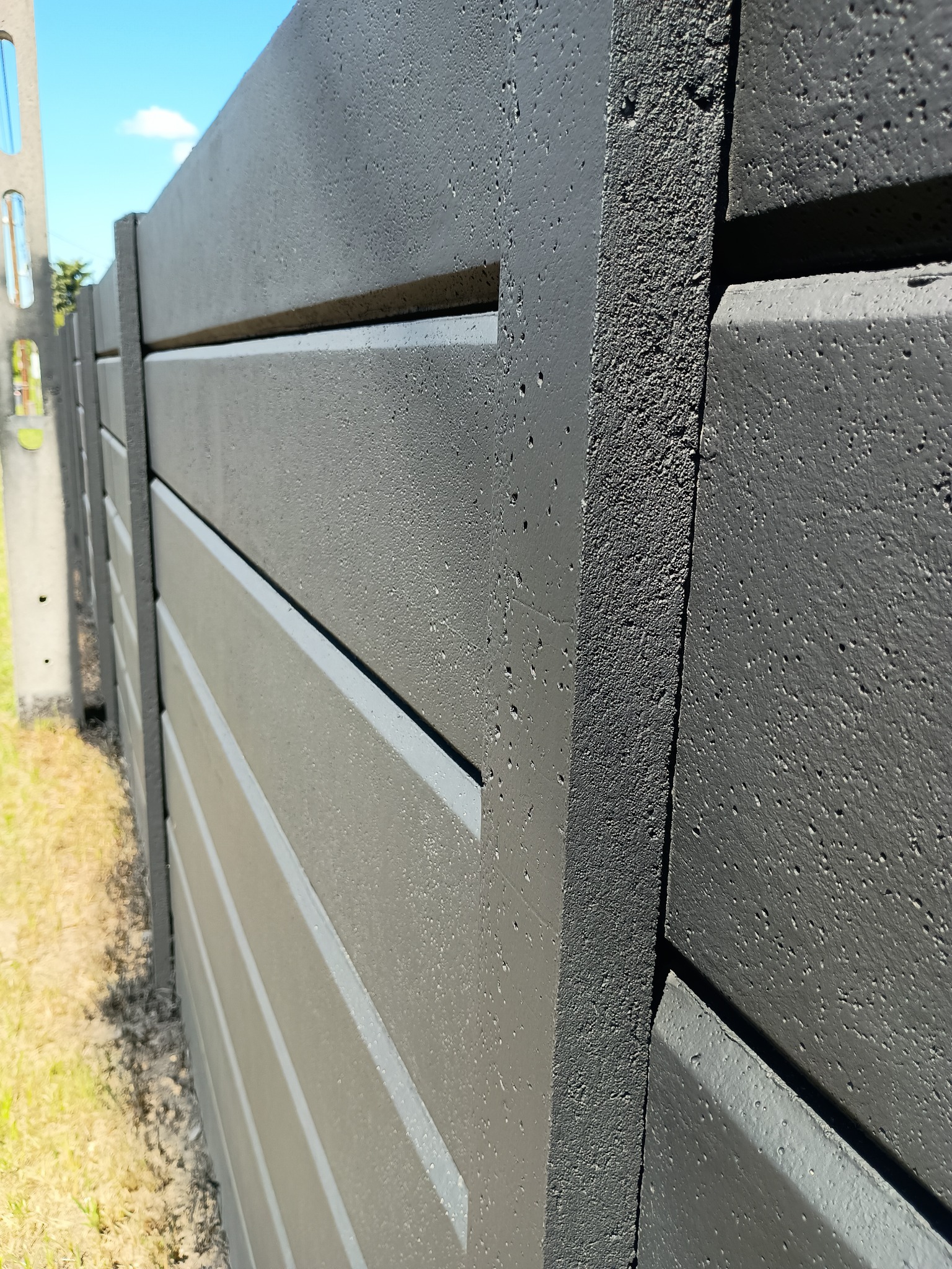 Szary, betonowy płot panelowy z poziomymi szczelinami, świeżo pomalowany w Miasteczku Śląskim. Widok z bliska, ukazujący fakturę betonu i precyzję wykonania.