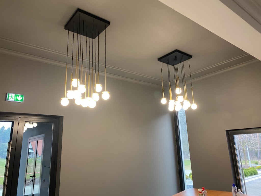 Dwa nowoczesne, czarne plafony z kulistymi, szklanymi kloszami, zawieszone w szarym pomieszczeniu z widocznym fragmentem stołu i oknami. Minimalistyczny design.