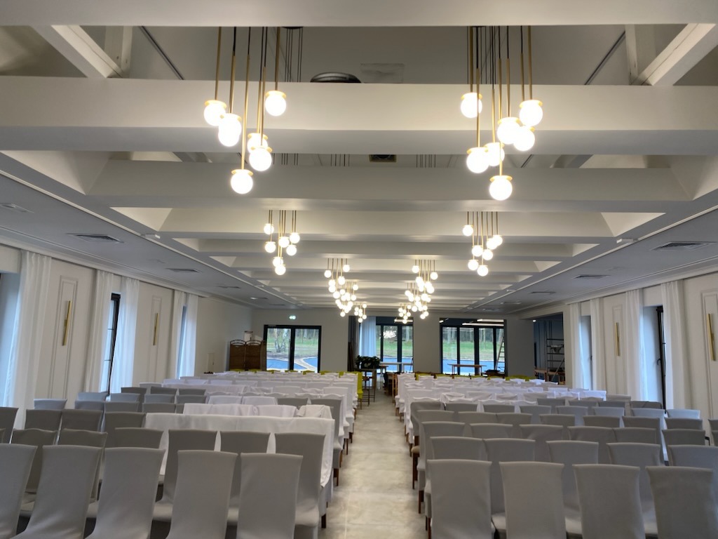 Jasne, przestronne wnętrze sali konferencyjnej z rzędami krzeseł w pokrowcach i nowoczesnym oświetleniem sufitowym. Widok na zewnątrz przez przeszklone drzwi.