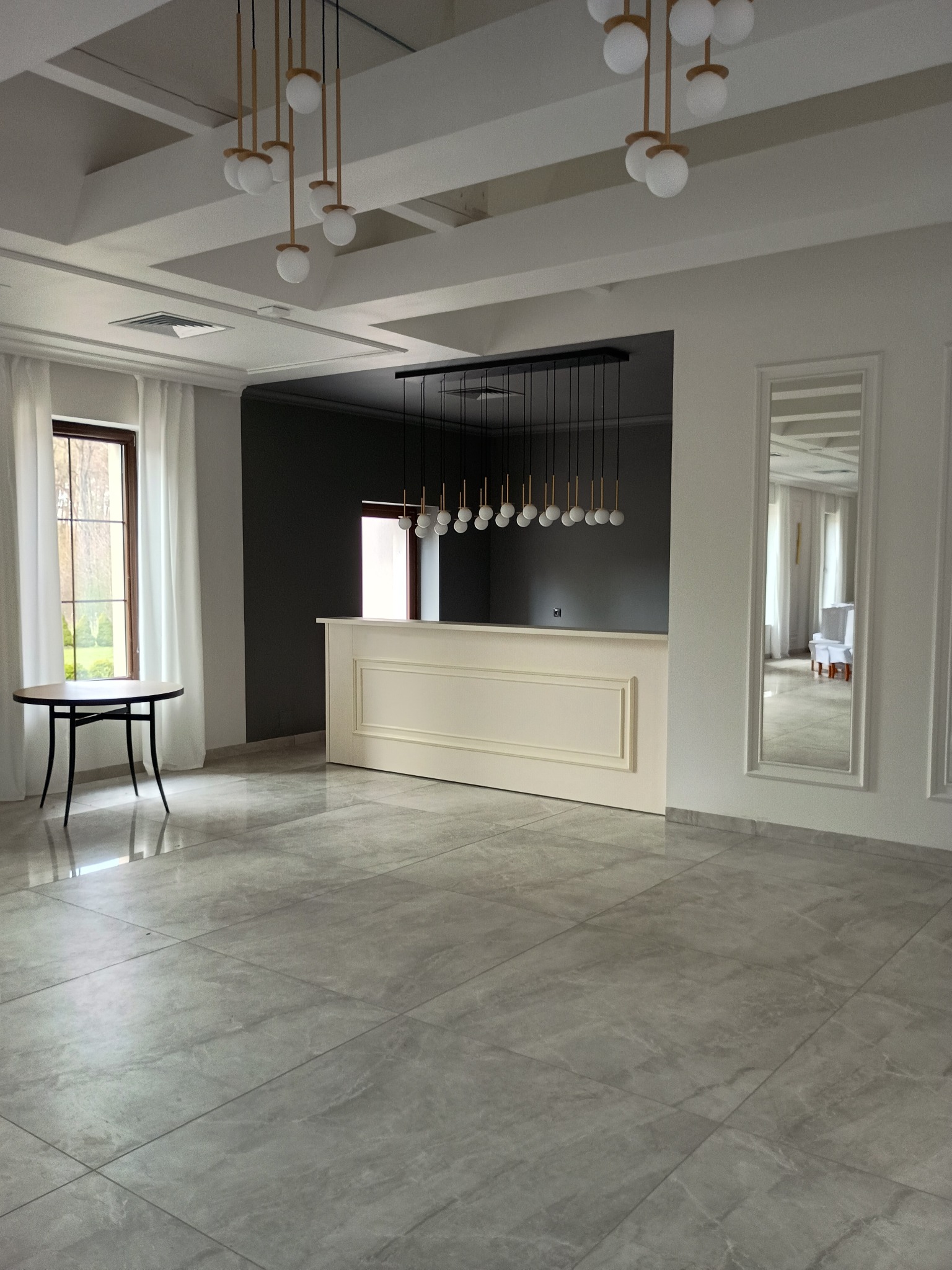 Eleganckie wnętrze po remoncie: szare płytki podłogowe, białe ściany z lustrami, designerskie lampy wiszące. Widok na salę z oknem i stolikiem.