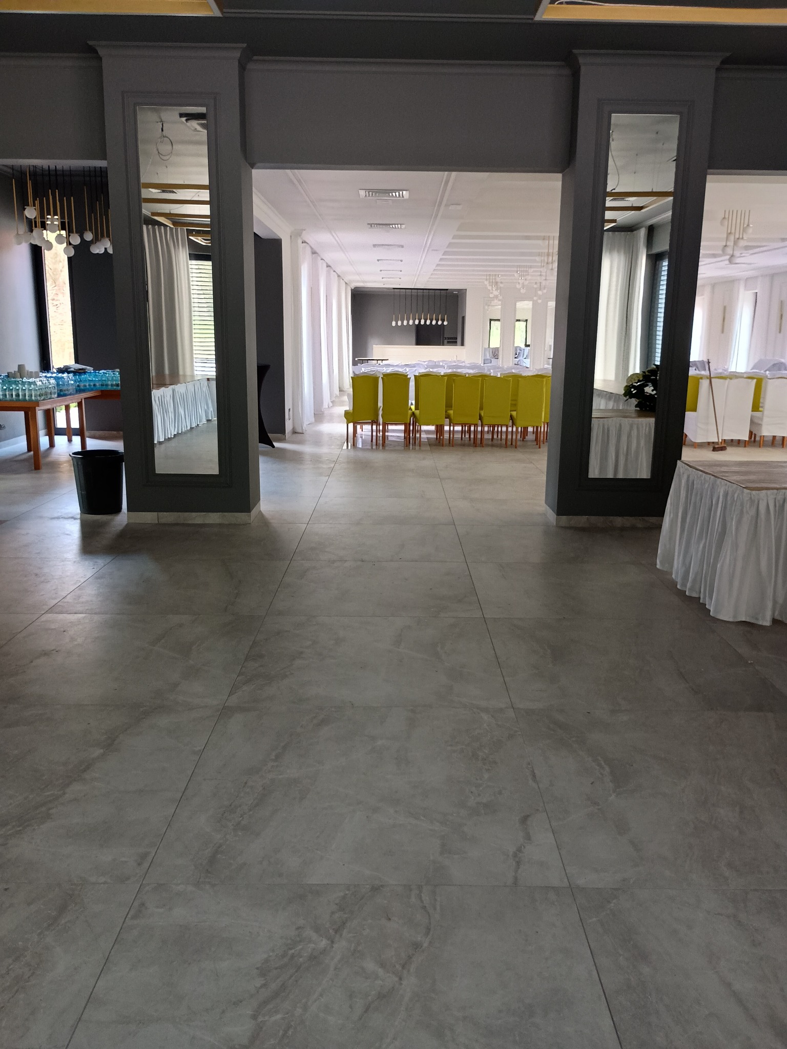 Sala bankietowa z szarymi płytkami podłogowymi, kolumnami z lustrami i rzędem żółtych krzeseł w tle. Widok na przygotowane stoły z białymi obrusami.