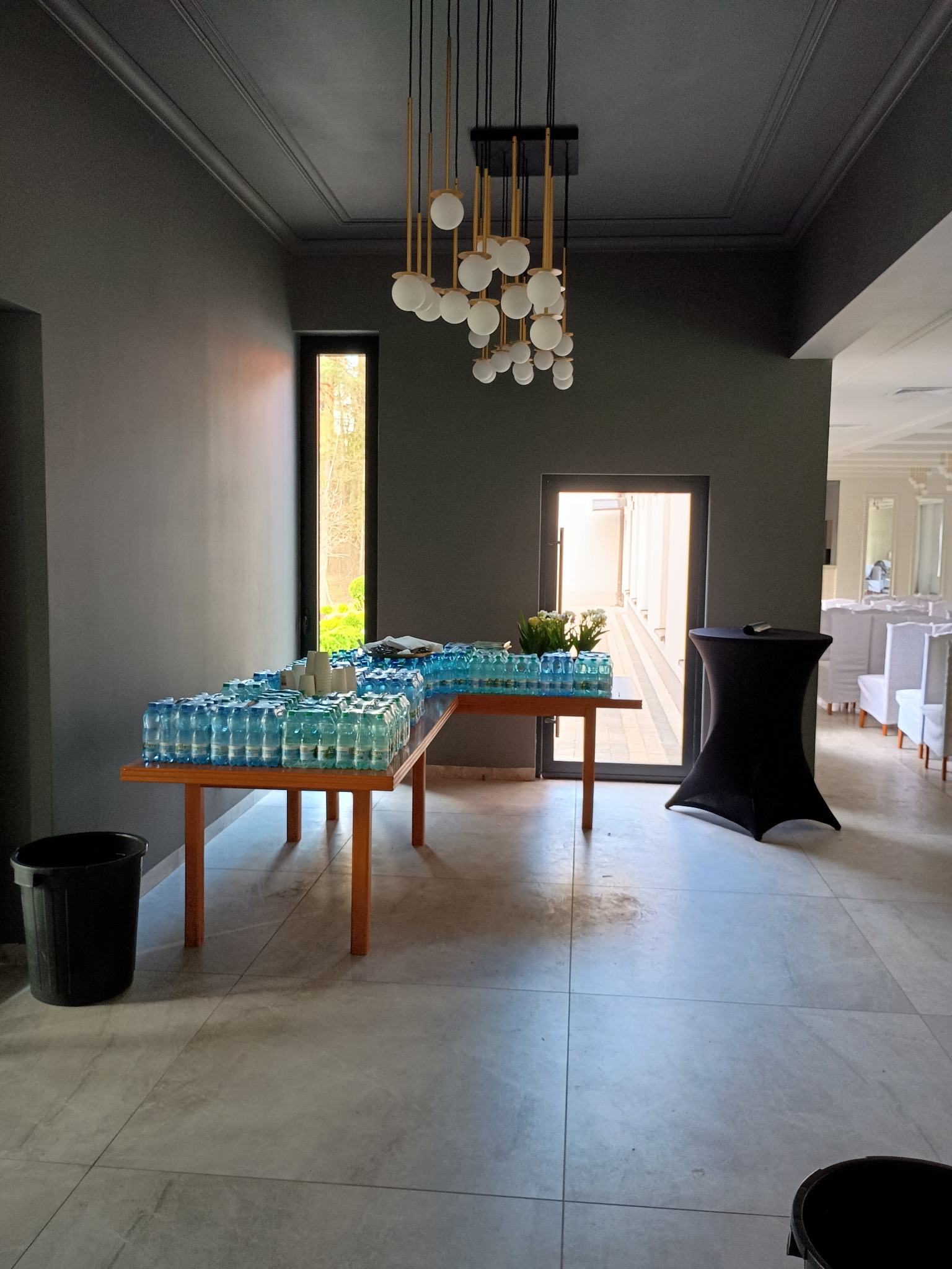 Eleganckie wnętrze z szarymi ścianami i sufitem, oświetlone designerską lampą. Na stole ułożone butelki wody, w tle widoczne krzesła i dekoracje, sugerujące przygotowania do wydarzenia.