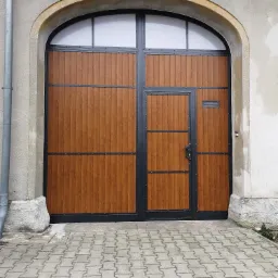 Dwuskrzydłowa brama garażowa z furtką, wykonana z paneli imitujących drewno, z czarnymi metalowymi ramami i elementami dekoracyjnymi, umieszczona w łukowatym otworze budynku.