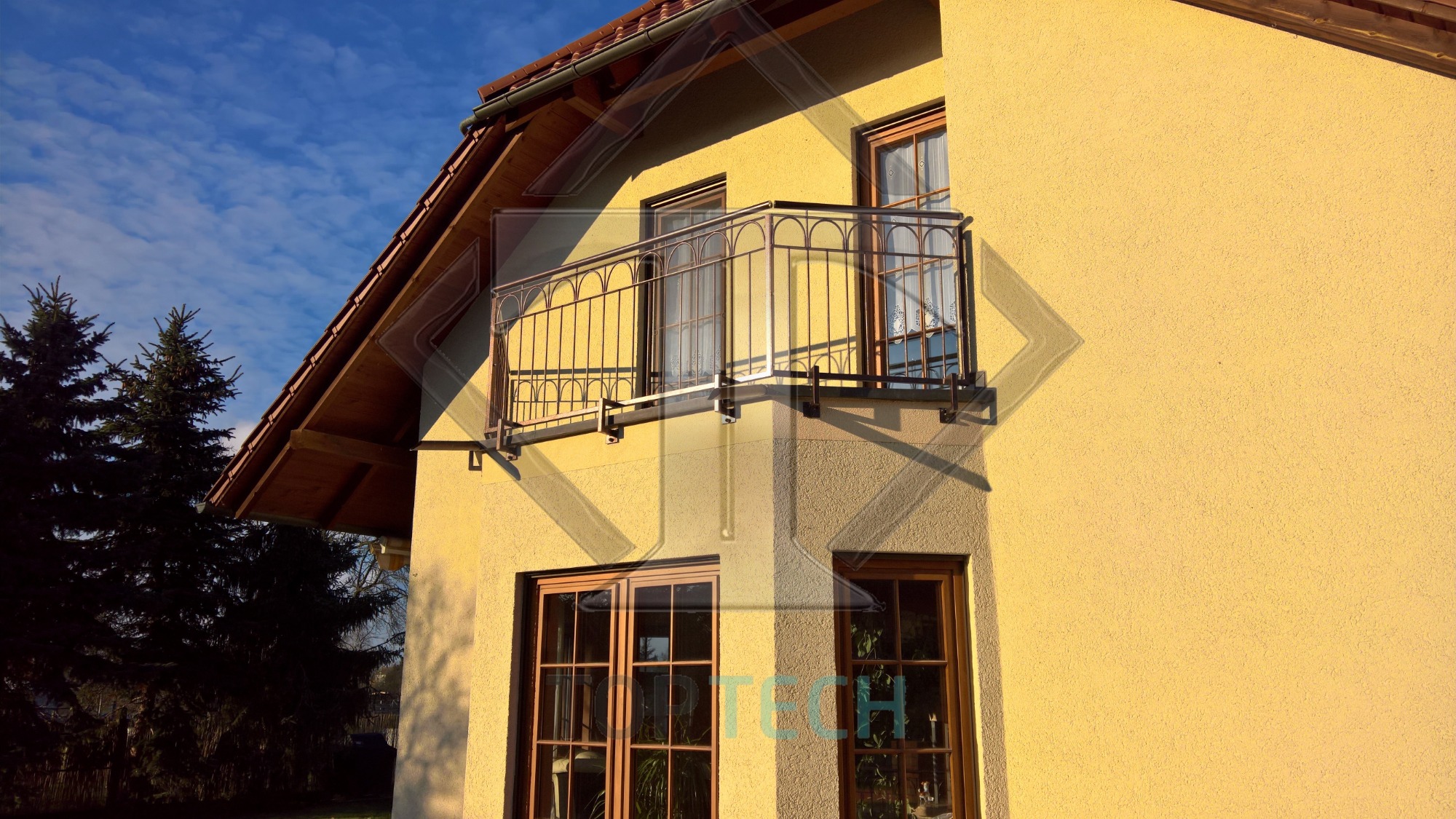 Kuta balustrada balkonowa z ozdobnymi elementami zamontowana na elewacji domu o żółtej barwie, widok z dołu na tle błękitnego nieba.