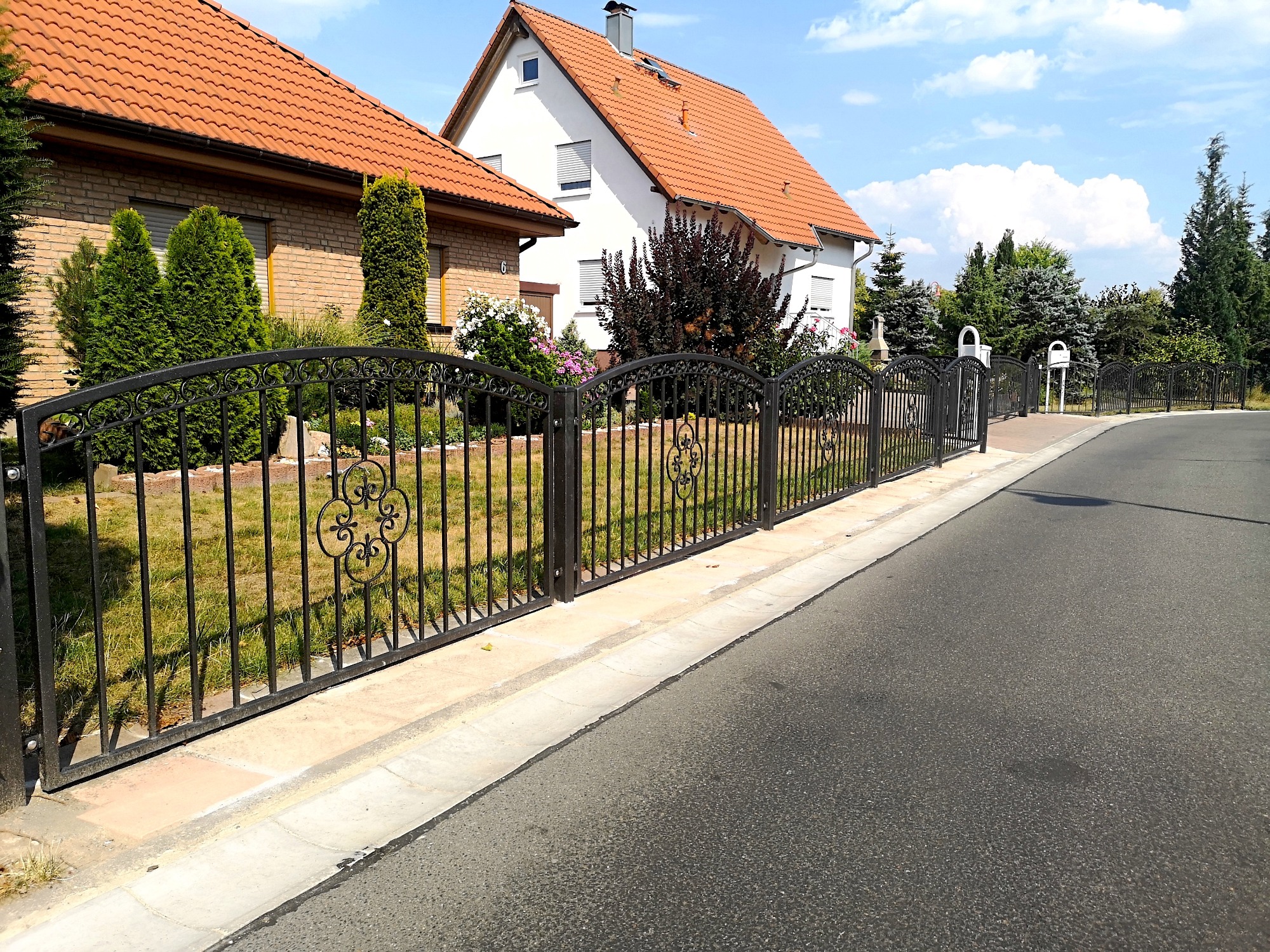 Eleganckie, czarne ogrodzenie z kutymi elementami zdobiące posesję z dwoma domami o czerwonych dachach, widoczne z poziomu asfaltowej drogi.