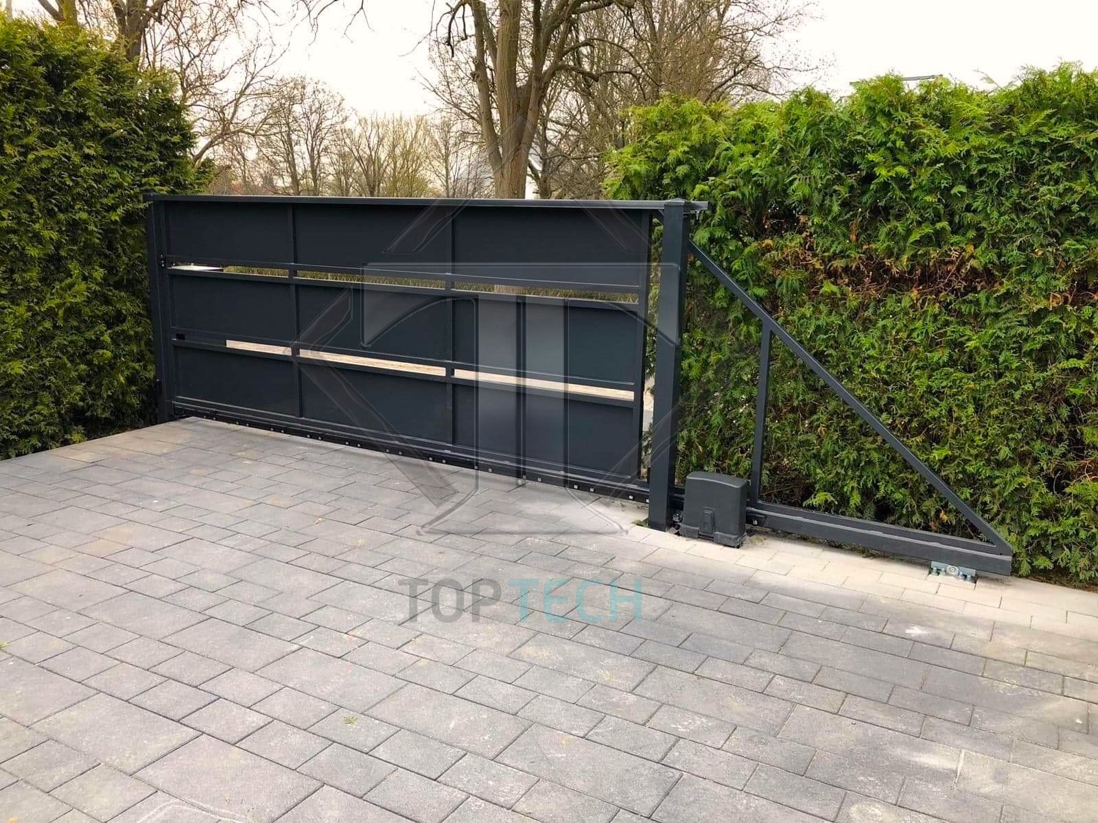 Automatyczna brama przesuwna w kolorze grafitowym, częściowo otwarta, z widocznym napędem i prowadnicą, na tle żywopłotu z tui.