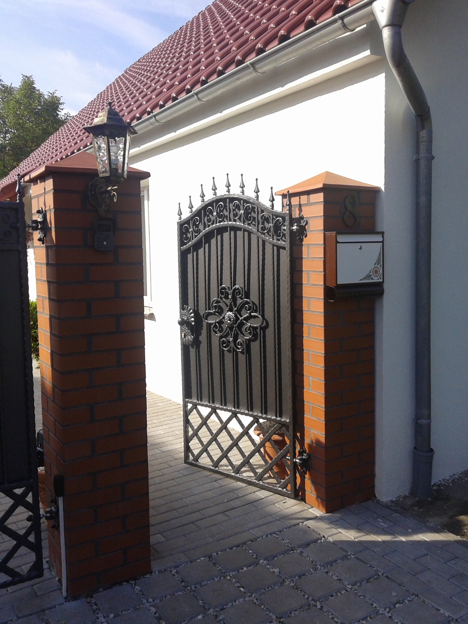 Otwarta, czarna, metalowa furtka z dekoracyjnymi elementami, umieszczona pomiędzy dwoma murowanymi słupkami z czerwonej cegły, na jednym ze słupków skrzynka pocztowa z numerem domu.
