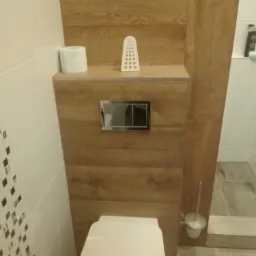 Nowoczesna toaleta podwieszana z drewnopodobną obudową, chromowanym przyciskiem spłukującym i ceramiczną szczotką toaletową, uzupełniona o dekoracyjny ażurowy element i rolkę papieru toaletowego.