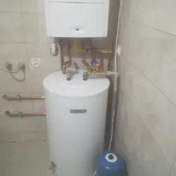 Bojler elektryczny zamontowany w łazience z podłączonymi rurami miedzianymi i zaworami, widoczny także mały zbiornik wyrównawczy koloru niebieskiego.