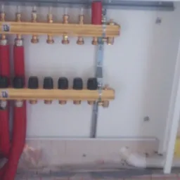 Rozdzielacz ogrzewania podłogowego z czerwonymi rurami w białej szafce, widoczne czarne pokrętła i mosiężne złączki, w tle biała ściana.