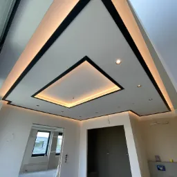 Sufit podwieszany z czarnymi obramowaniami i oświetleniem LED, widok z dołu na surowy stan wnętrza z widocznymi przewodami i otworami drzwiowymi.