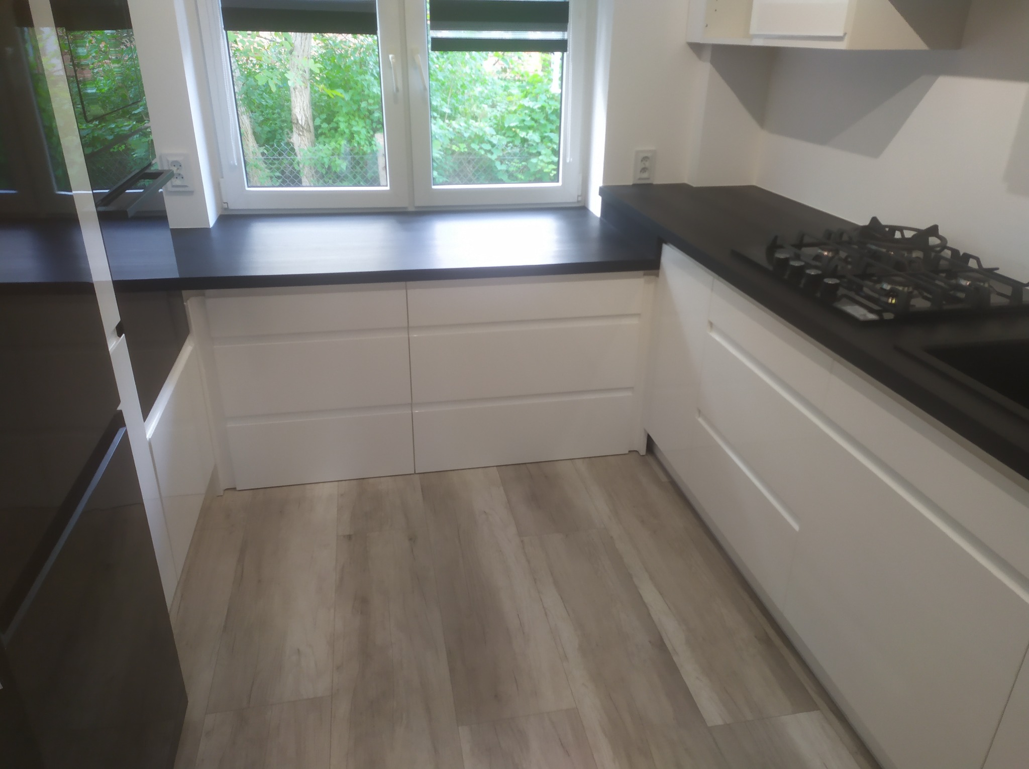 Nowoczesna, jasna kuchnia z białymi szafkami i czarnym blatem, widok na okno z zielenią. Na blacie kuchenka gazowa. Podłoga wyłożona panelami imitującymi drewno.