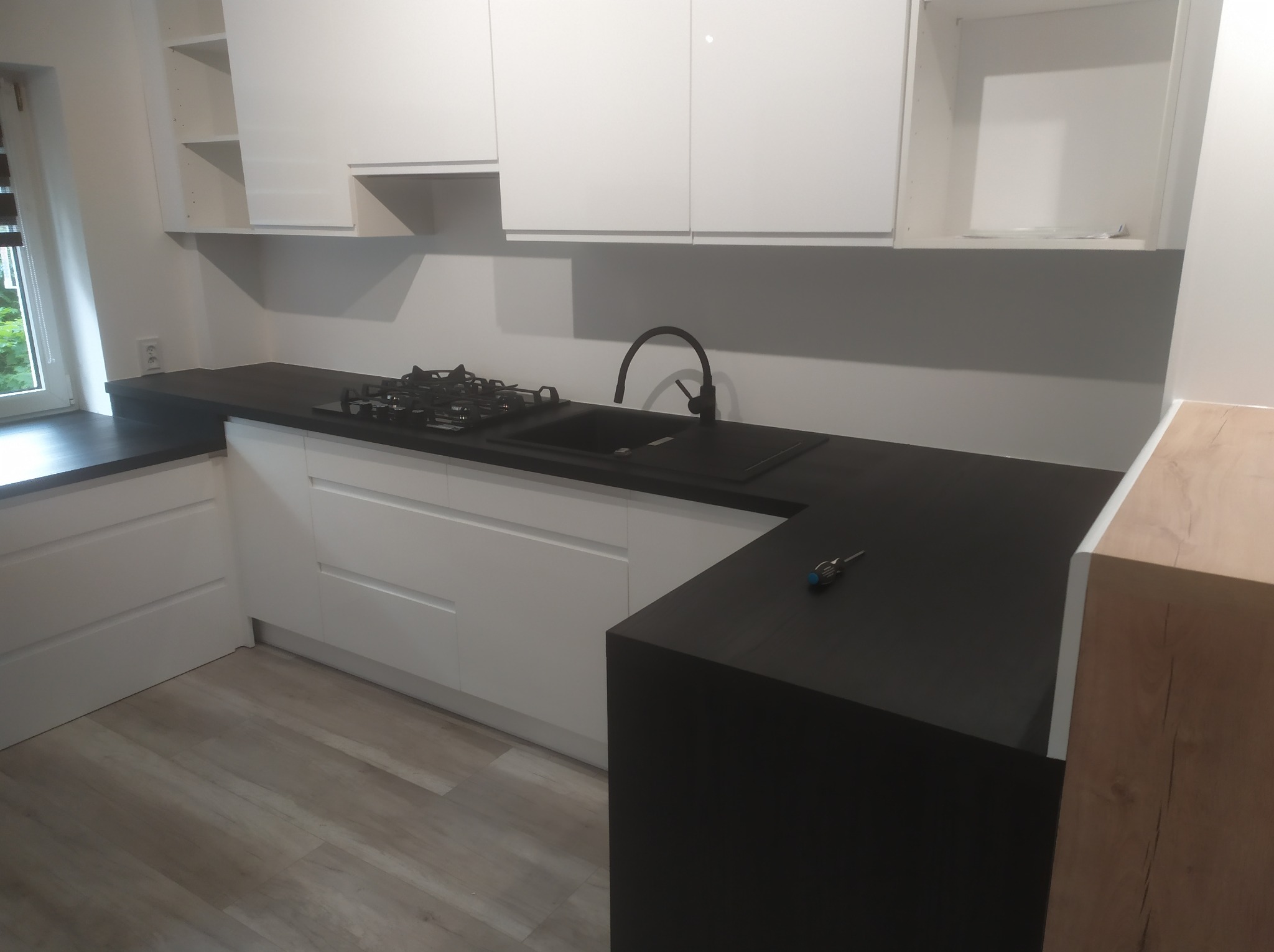 Nowoczesna kuchnia po remoncie: białe szafki, czarny blat, płyta gazowa i zlew. Na blacie leży śrubokręt. Podłoga wyłożona panelami.