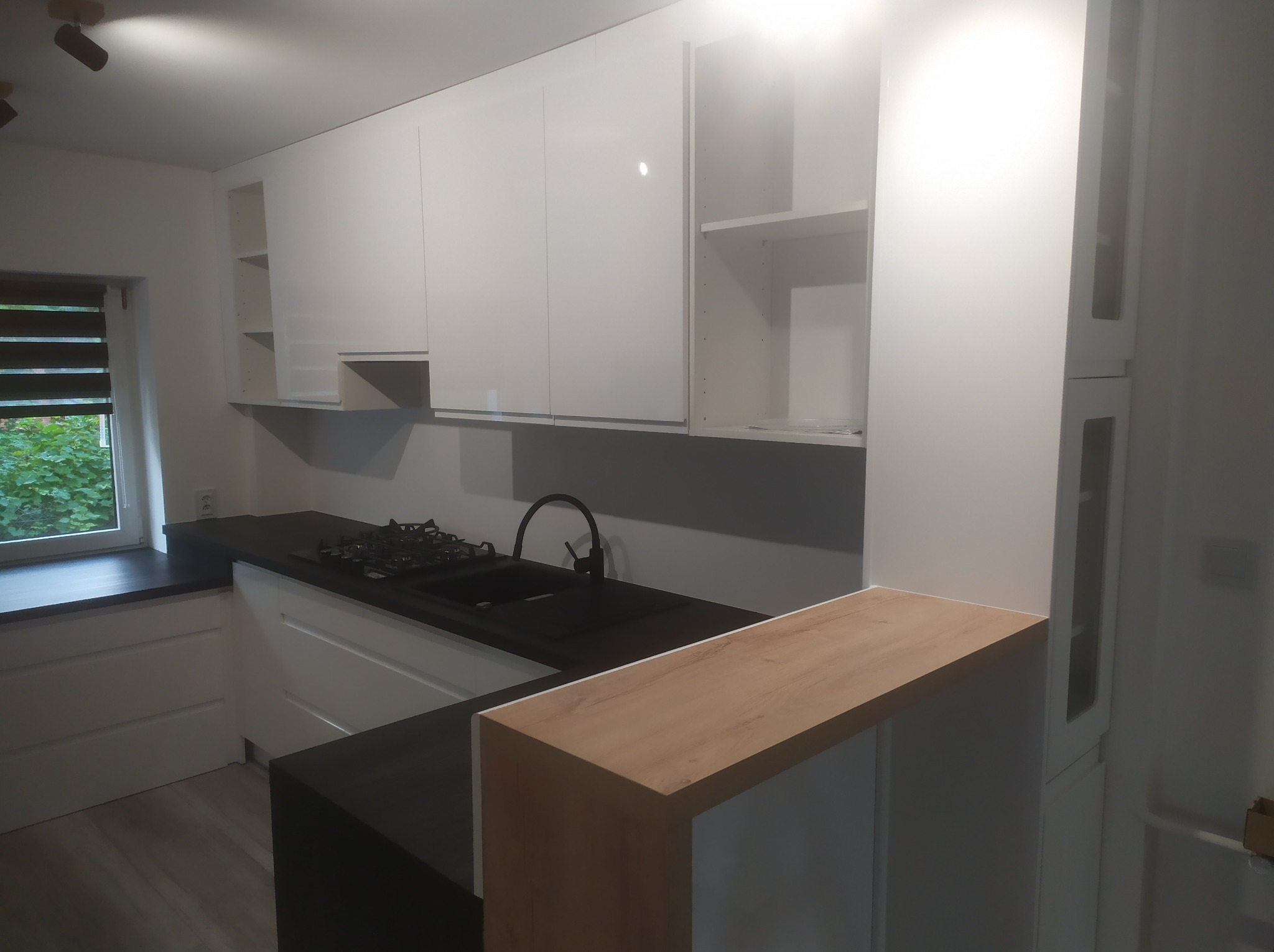 Nowoczesna, jasna kuchnia z białymi szafkami i czarnym blatem. Widoczny fragment okna i drewniany element barku. Oświetlenie punktowe.