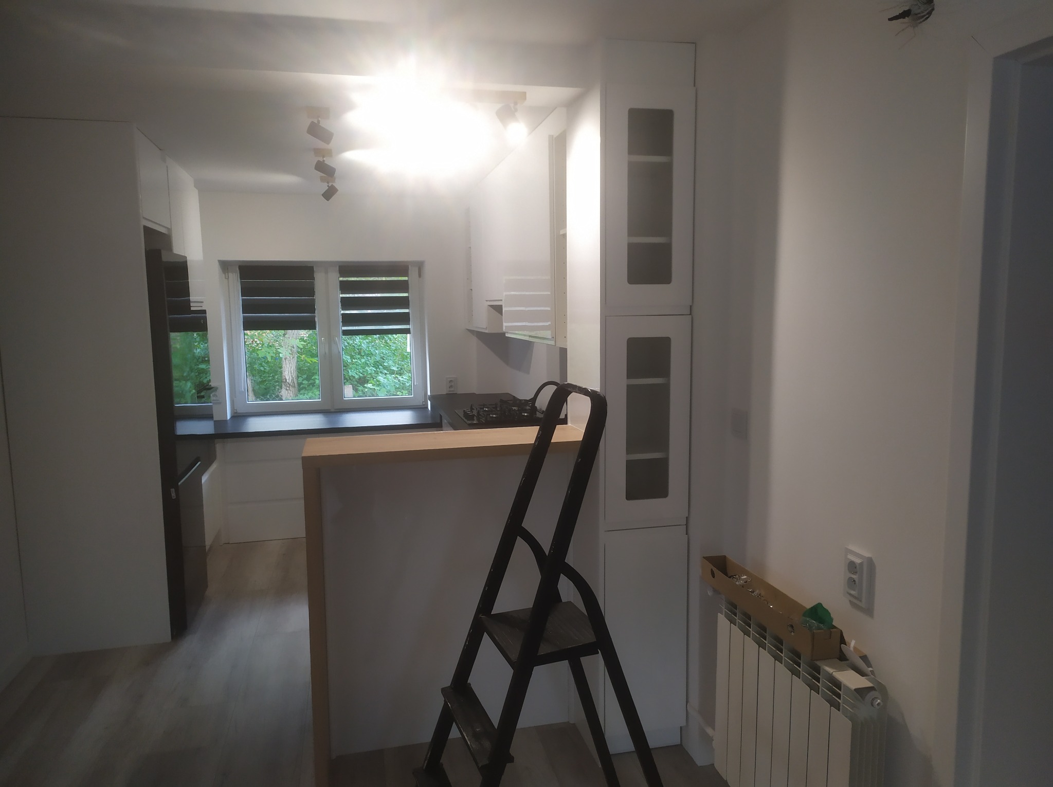 Jasna kuchnia w trakcie prac wykończeniowych: białe szafki, drewniany blat i drabina malarska w centrum. Oświetlenie sufitowe i okno z widokiem na zieleń.