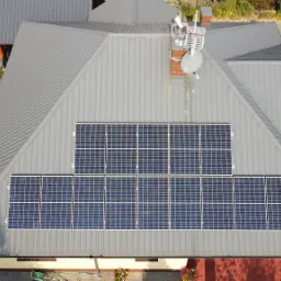 Widok z drona na szary dach z panelami słonecznymi, kominem z anteną i satelitą, oraz tunelem foliowym w ogrodzie.