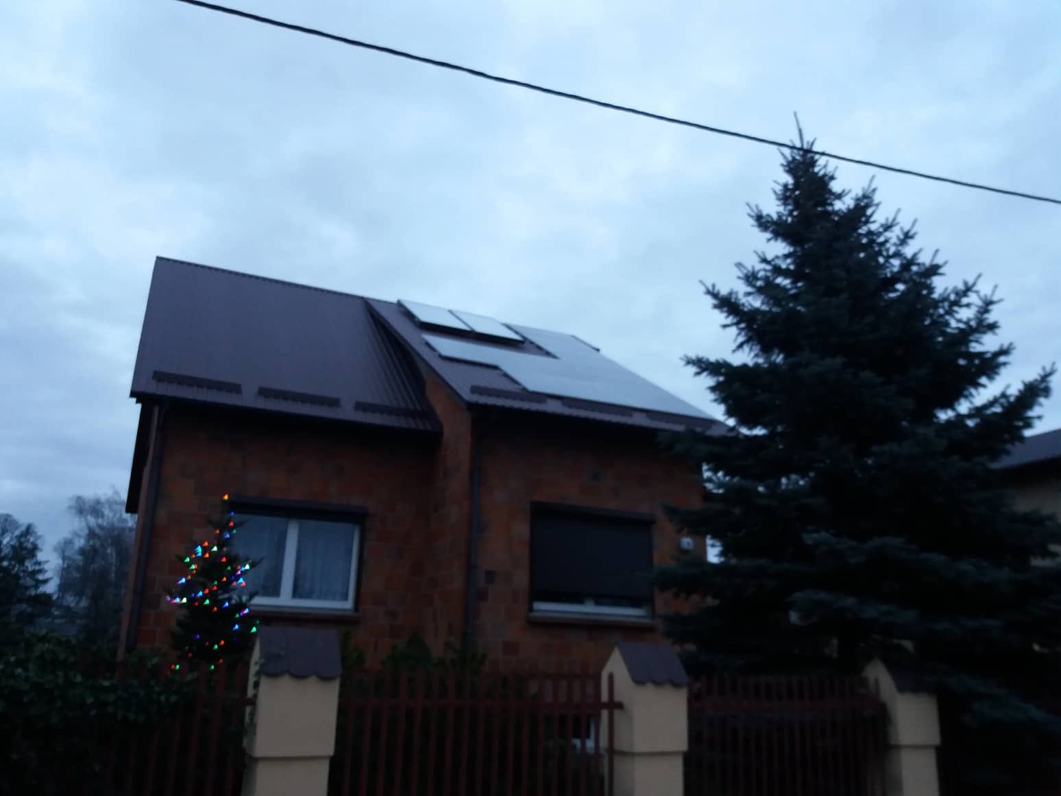 Dom jednorodzinny z brązowym dachem i panelami słonecznymi na dachu, obok choinka z kolorowymi lampkami, pochmurne niebo.