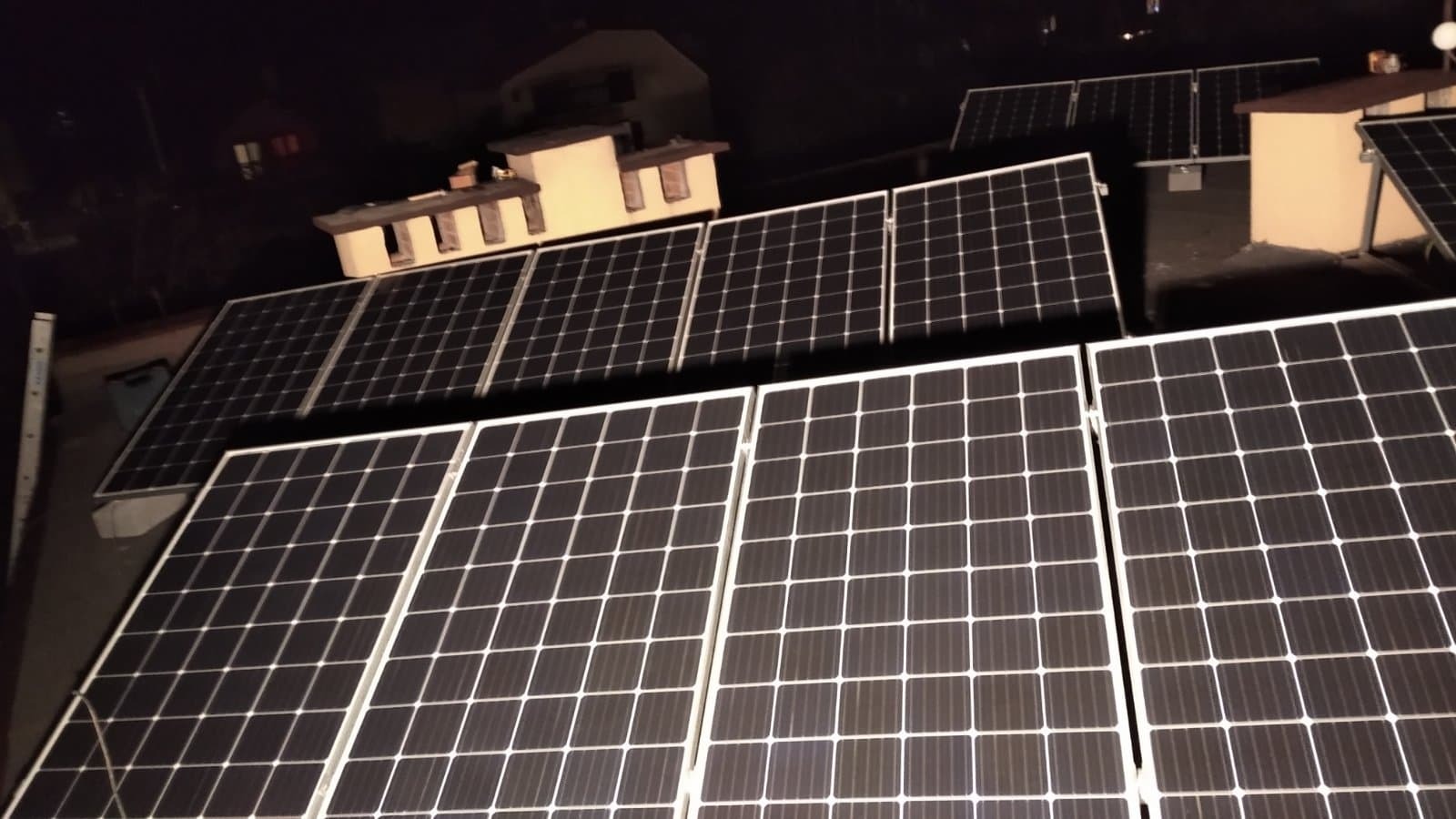 Panele fotowoltaiczne na dachu budynku oświetlone nocnym światłem, widoczne kominy wentylacyjne.