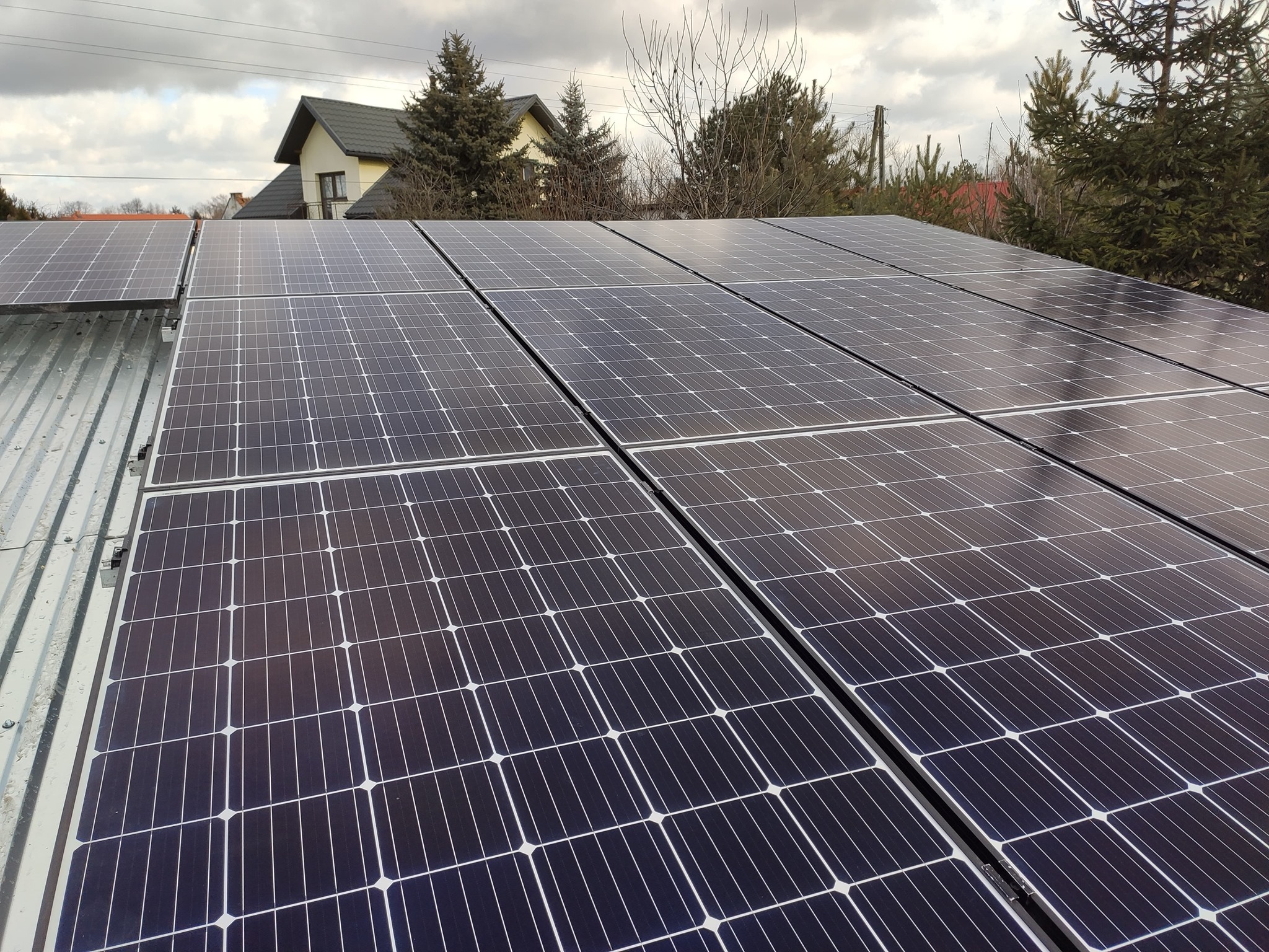Rząd paneli słonecznych na metalowym dachu domu jednorodzinnego w pochmurny dzień. Widoczne drzewa i budynek w tle.