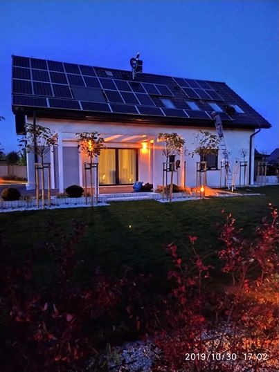 Dom jednorodzinny z panelami fotowoltaicznymi na dachu, widoczna drabina oparta o ścianę, oświetlenie zewnętrzne włączone, zdjęcie wykonane o zmierzchu.