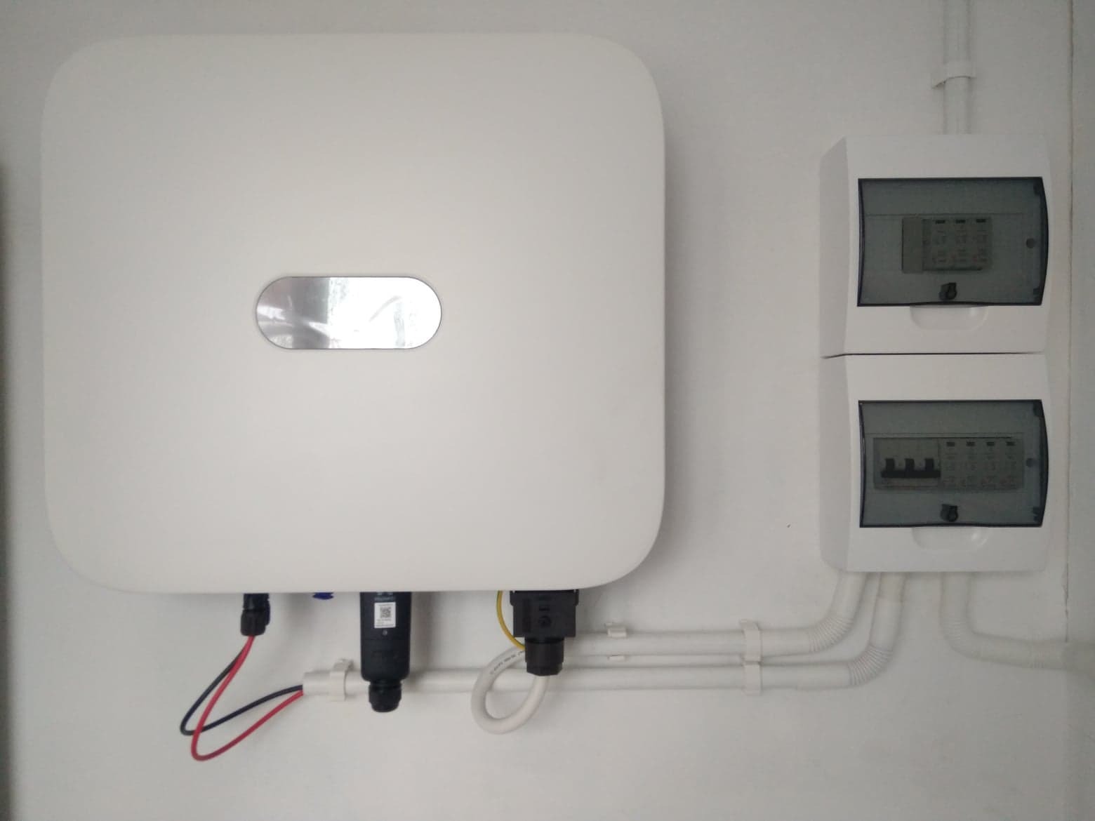 Biały falownik zamontowany na ścianie, z widocznymi przewodami i dwoma skrzynkami zabezpieczeń.