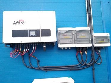 Biały falownik firmy Afore zamontowany na niebieskiej ścianie, z podłączonymi kablami i trzema skrzynkami rozdzielczymi obok.