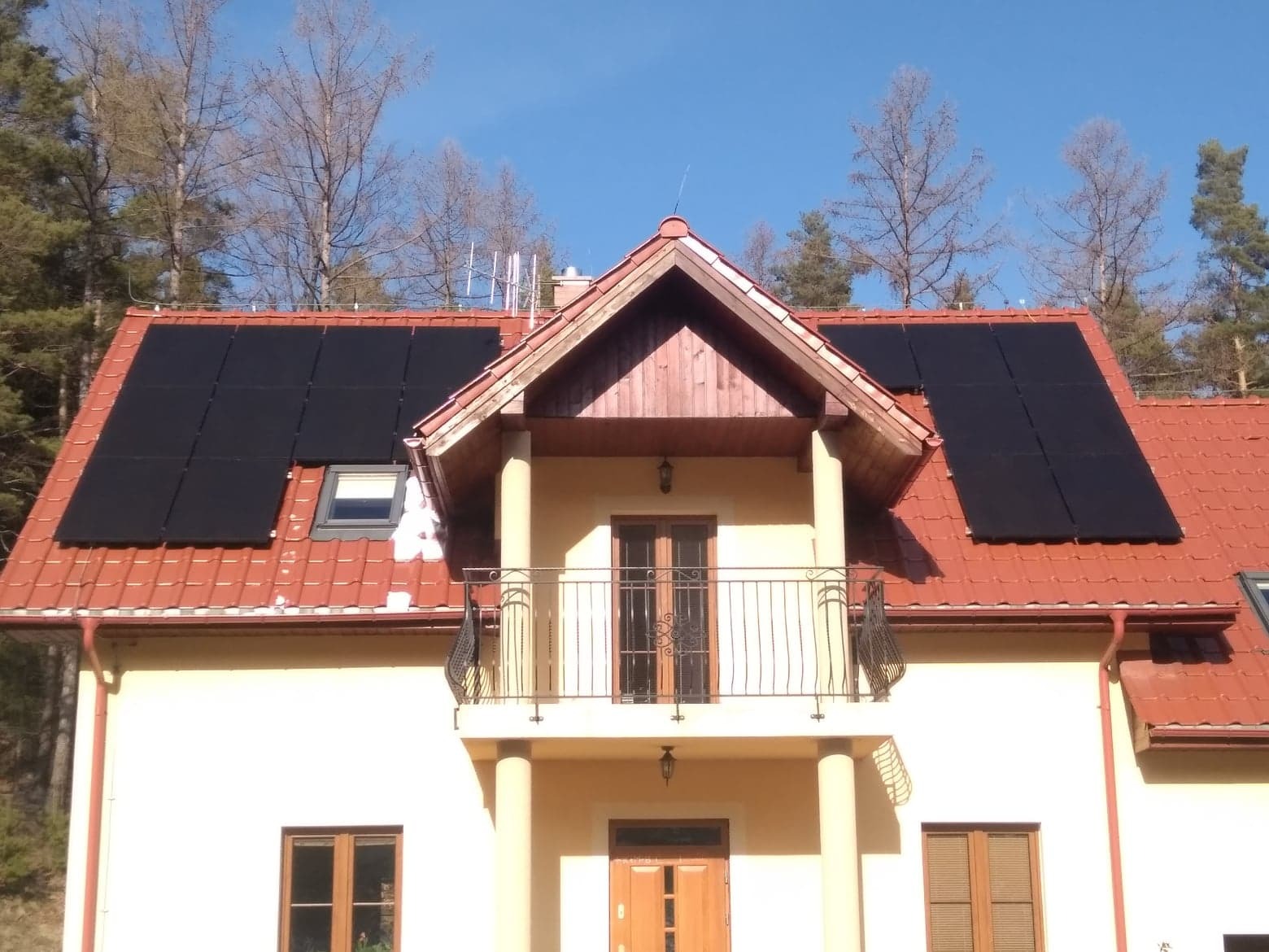 Dom jednorodzinny z czerwoną dachówką, na której zamontowano panele fotowoltaiczne; widoczny balkon z metalową balustradą i drzewa w tle.