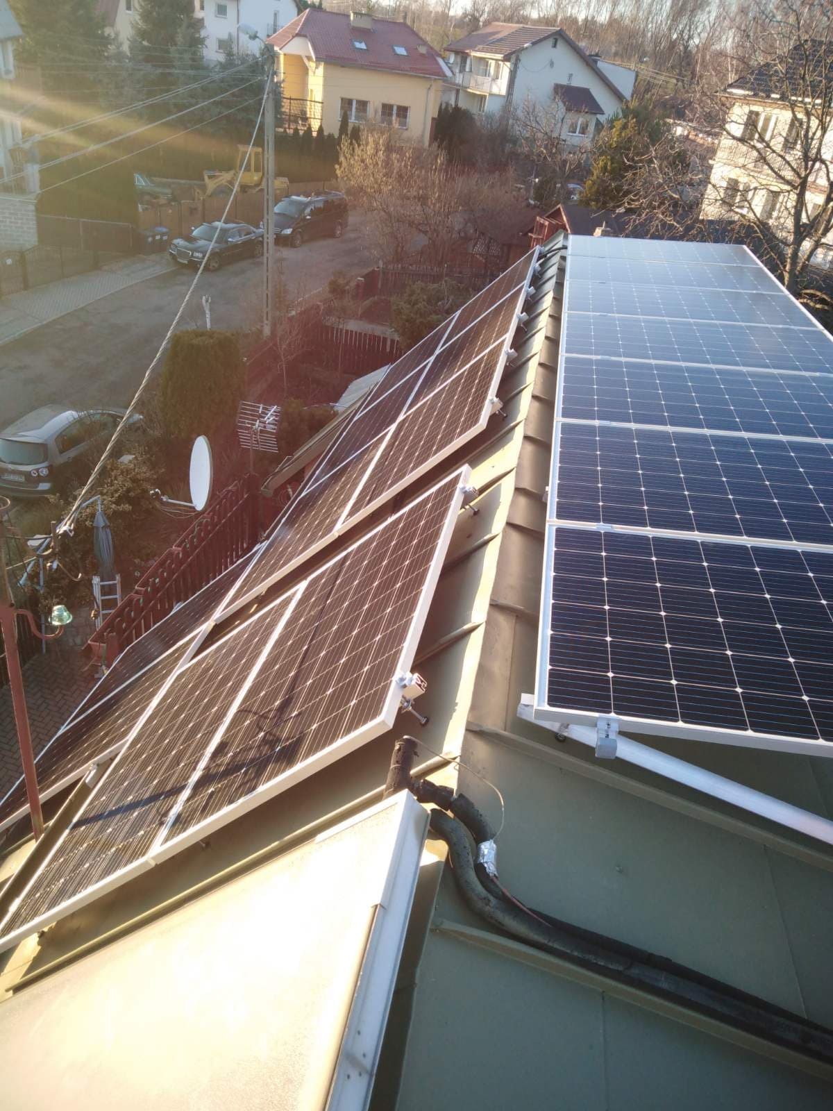 Panele słoneczne na dachu domu jednorodzinnego, widok z góry, w tle zabudowa mieszkalna i drzewa.