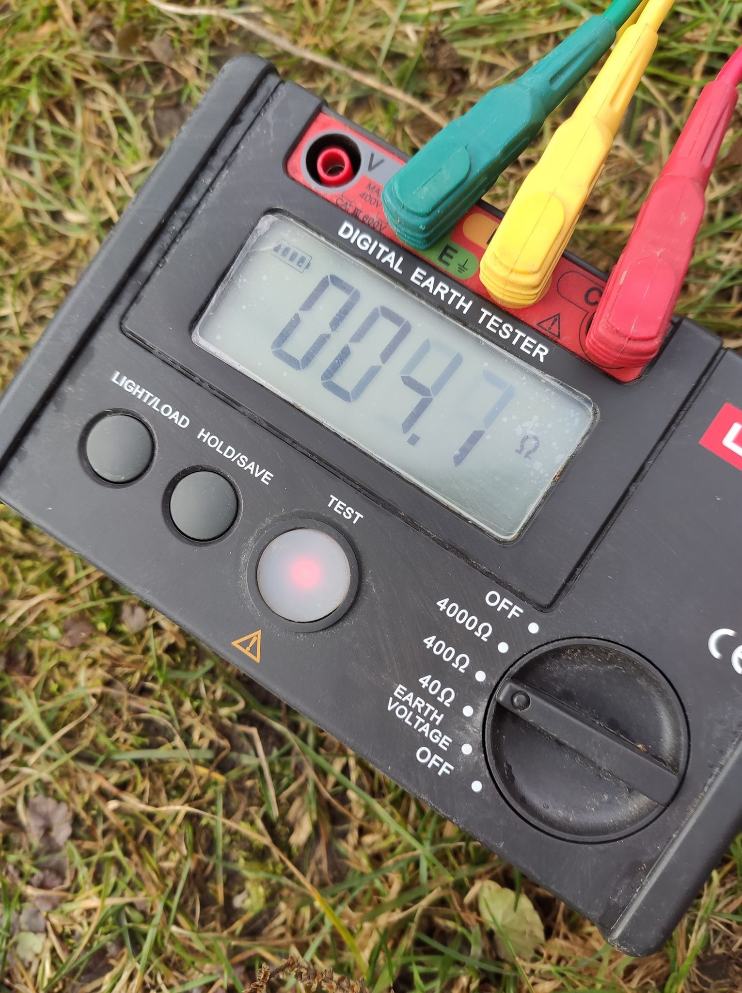 Cyfrowy miernik rezystancji uziemienia z podłączonymi przewodami pomiarowymi na tle trawy, wskazujący wartość 004.7 Ω.