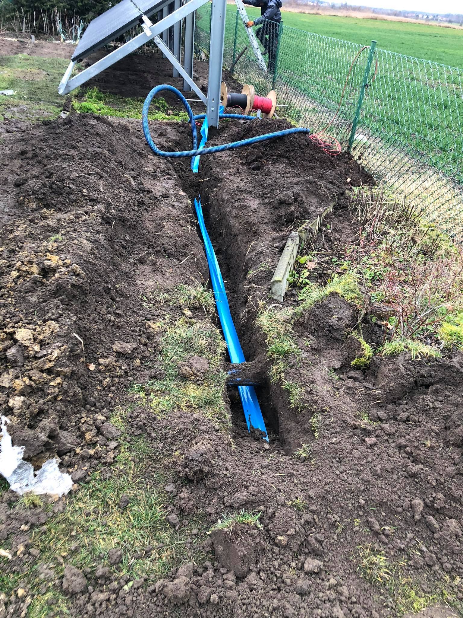 Wykopany rów w ziemi z niebieską rurą ochronną na kable, widoczny panel słoneczny na konstrukcji wsporczej, w tle osoba na drabinie przy ogrodzeniu z siatki.