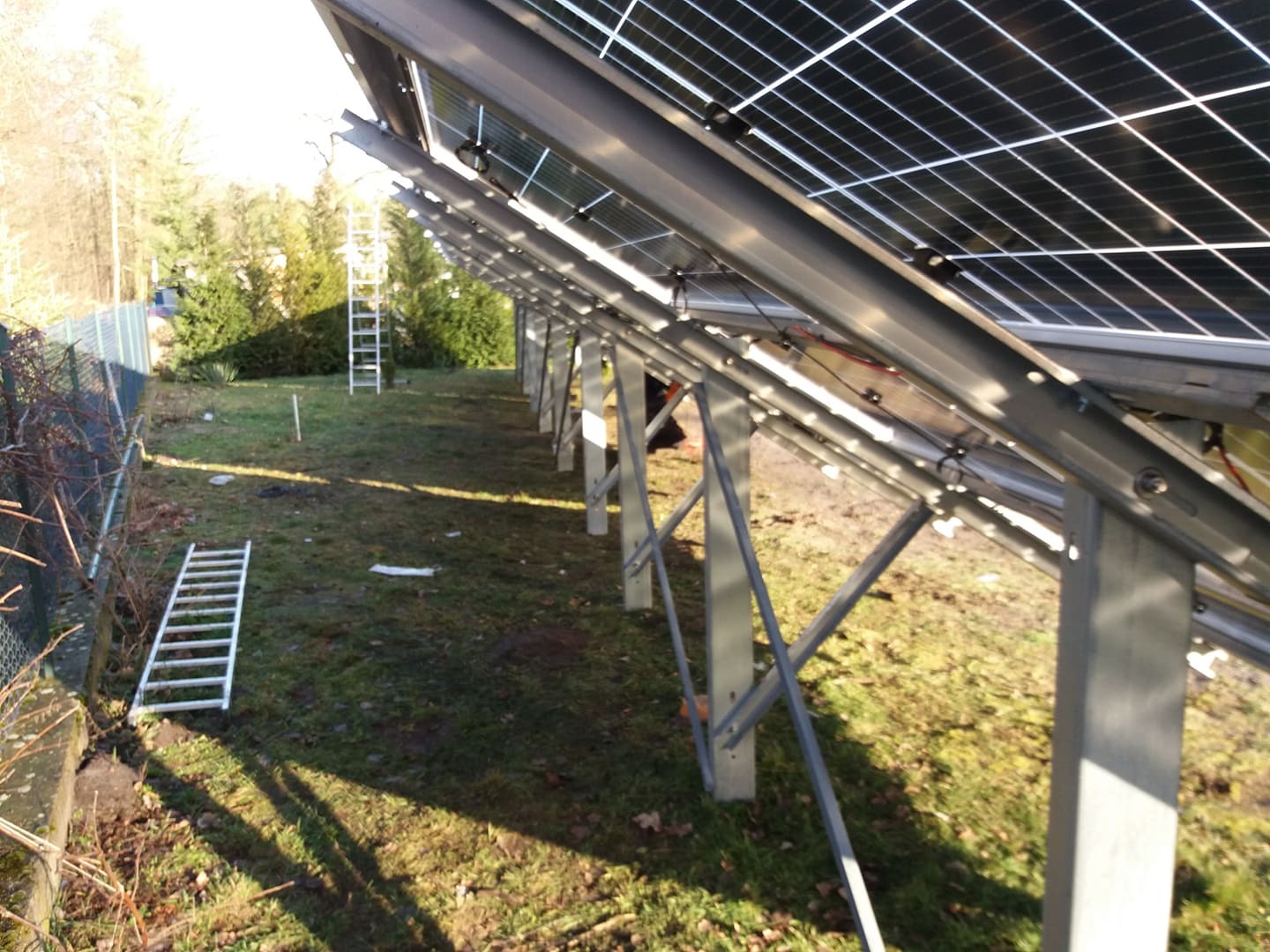 Montaż paneli na konstrukcji wsporczej na trawiastym terenie, widoczne dwie drabiny aluminiowe, częściowo zamontowane panele, słoneczny dzień.