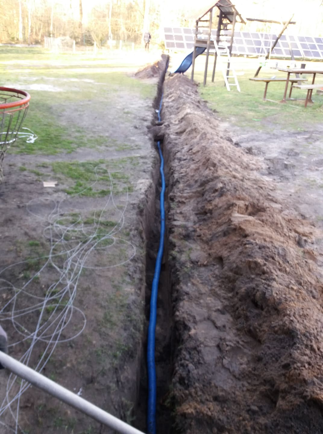 Wykop w ziemi z niebieską rurą wewnątrz, prawdopodobnie przygotowanie pod instalację kabli do paneli słonecznych widocznych w tle.