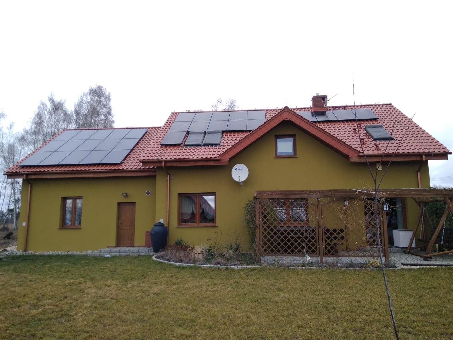 Dom jednorodzinny z panelami fotowoltaicznymi na dachu z czerwonej dachówki, widoczne okna dachowe i antena satelitarna, trawnik przed domem.