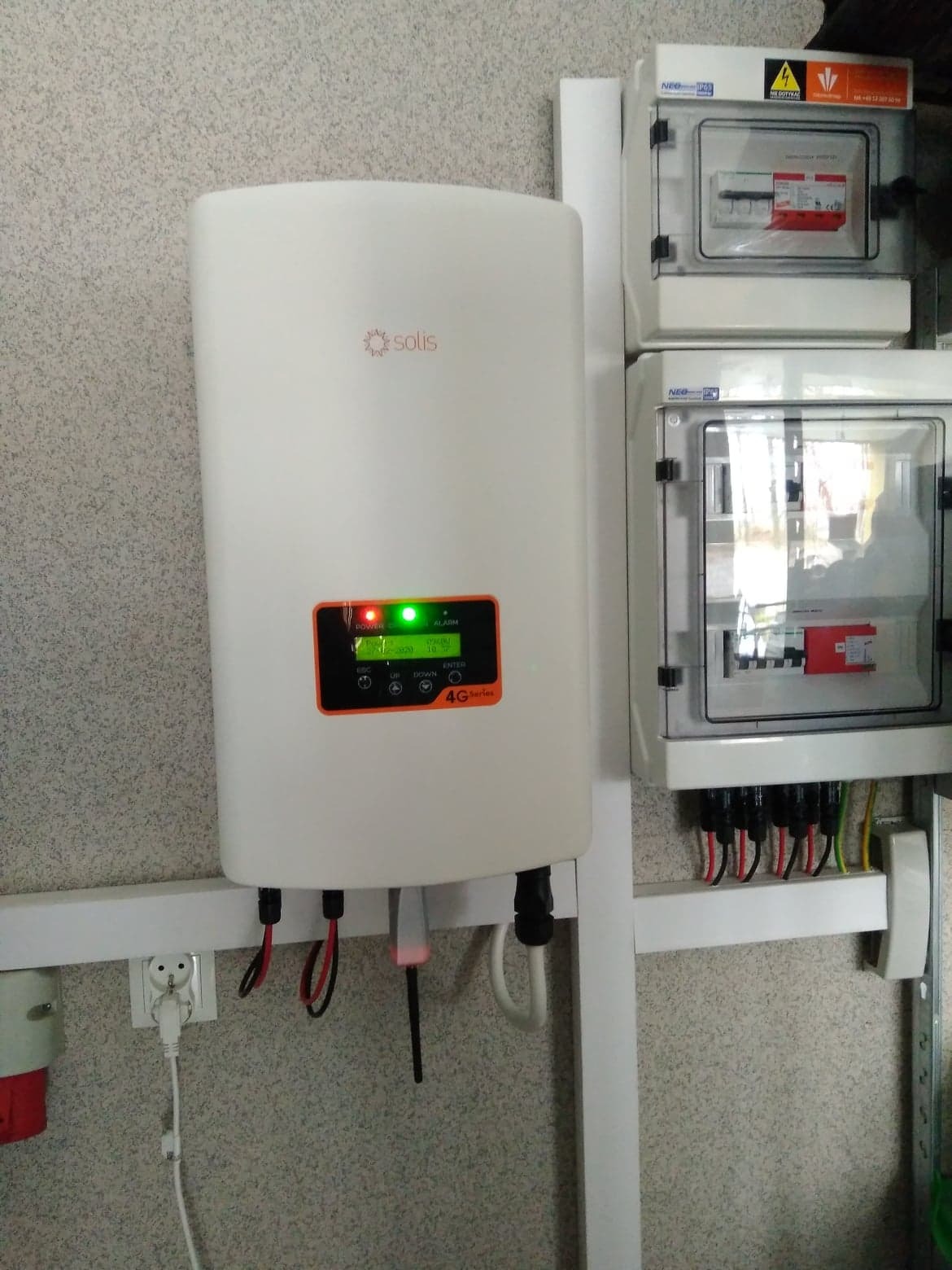 Biały inwerter Solis 4G Series zamontowany na ścianie obok dwóch skrzynek elektrycznych z widocznymi bezpiecznikami i przewodami.